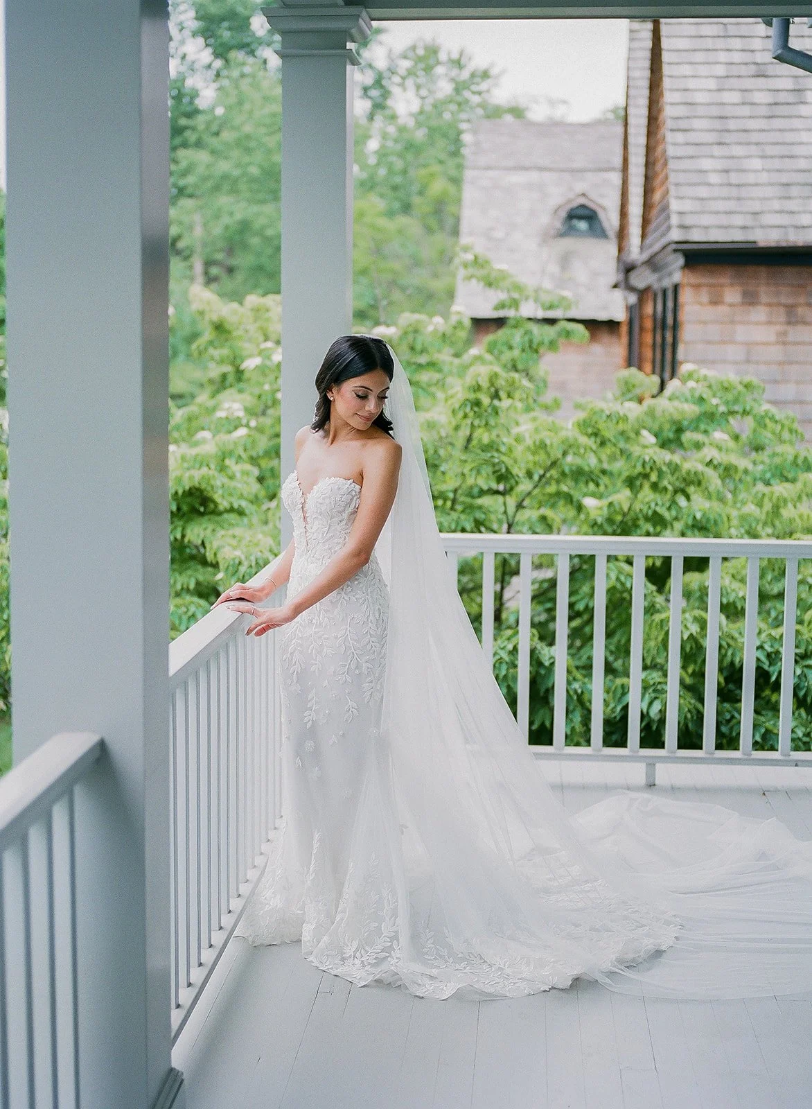 A bride in a strapless white wedding gown with lace details, standing on a porch with a white railing, looking down and to the side, with a long veil flowing behind her. Green trees and a brick house are visible in the background.