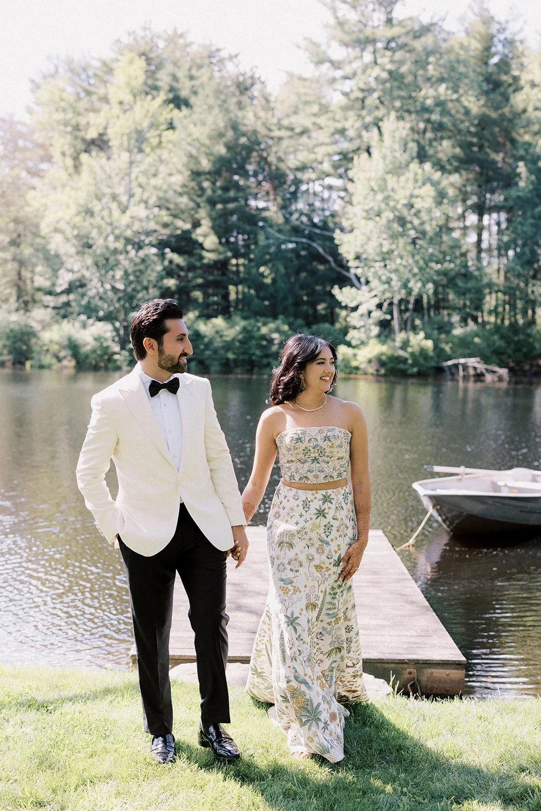 A couple dressed in formal attire walking hand in hand along a grassy area by a lake, with trees in the background and a small boat tied to a dock.