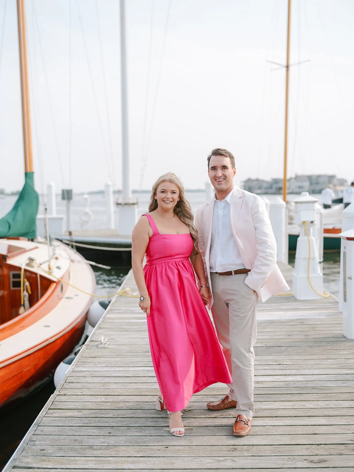 A couple standing on a wooden dock near sailboats, smiling and holding hands. The woman is wearing a bright pink dress and the man is in a beige suit with a white shirt.