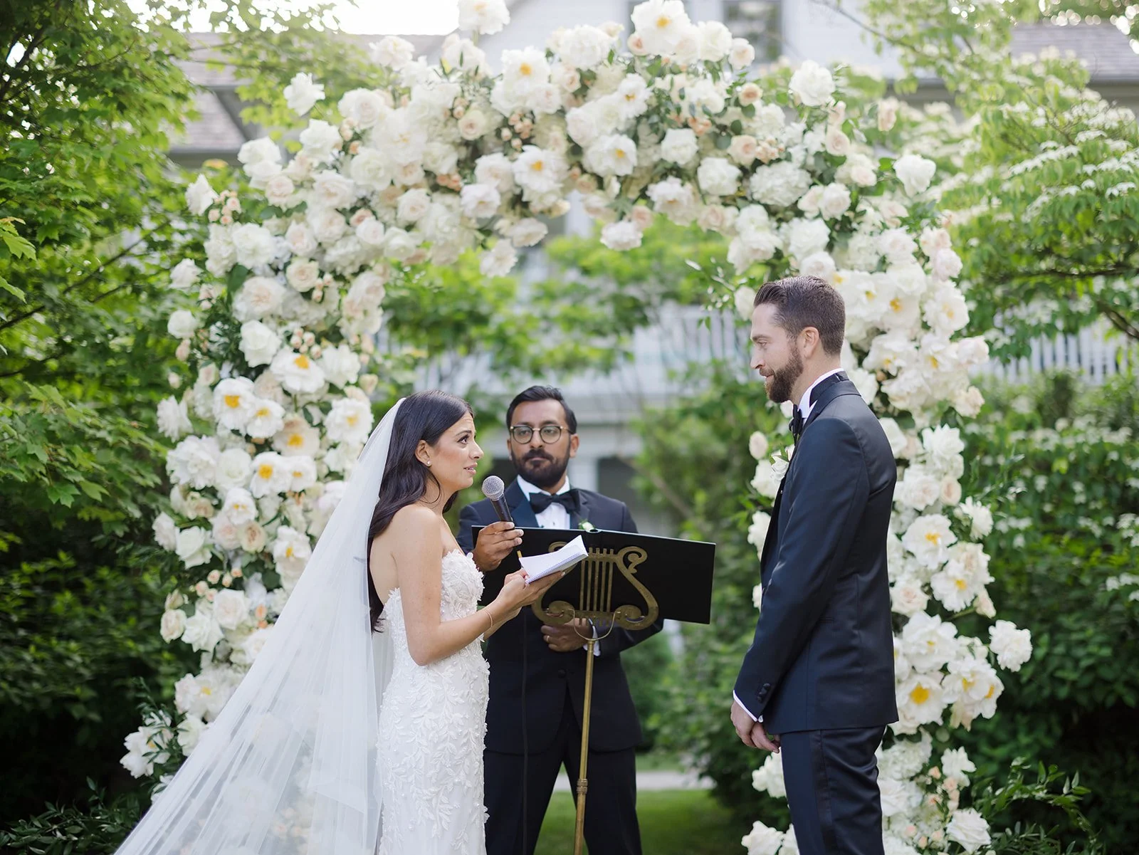 A bride and groom exchange vows during an outdoor wedding ceremony beneath a floral arch of white flowers, with an officiant standing between them holding a microphone and a book.