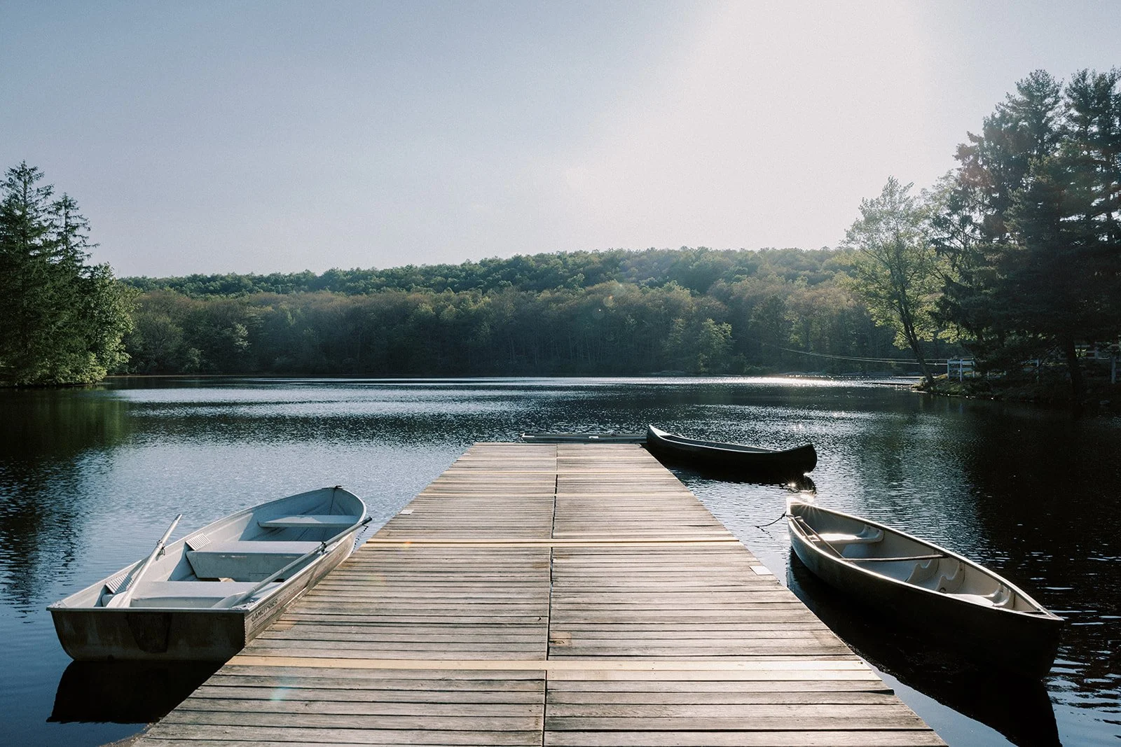 A wooden dock extending into a calm lake with three small boats, surrounded by trees and hills under a clear sky.