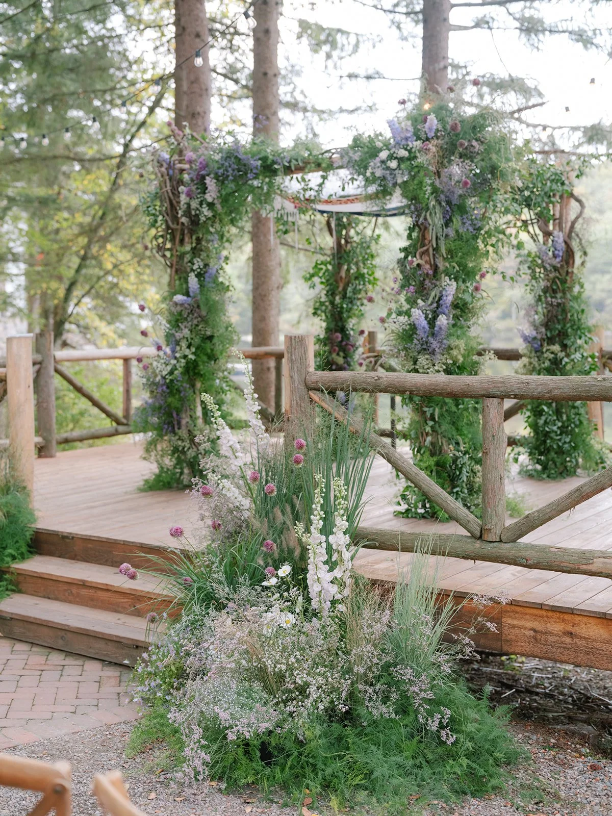 Wooden ceremony arch decorated with purple and white flowers, set in a forested outdoor setting with string lights overhead.
