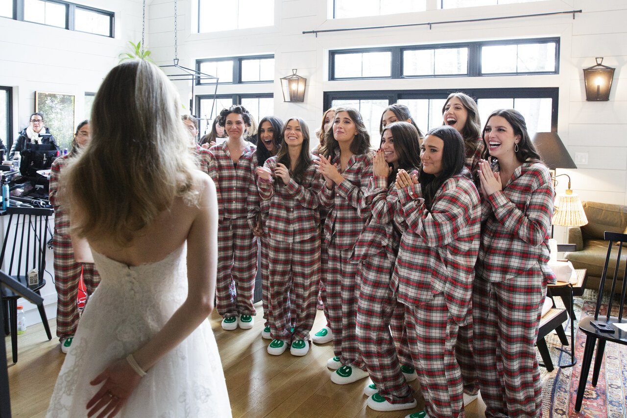 Bride in a white gown facing a group of women in matching plaid pajamas, some with hands clasped or covering their mouths, in a cozy, well-lit room with large windows.