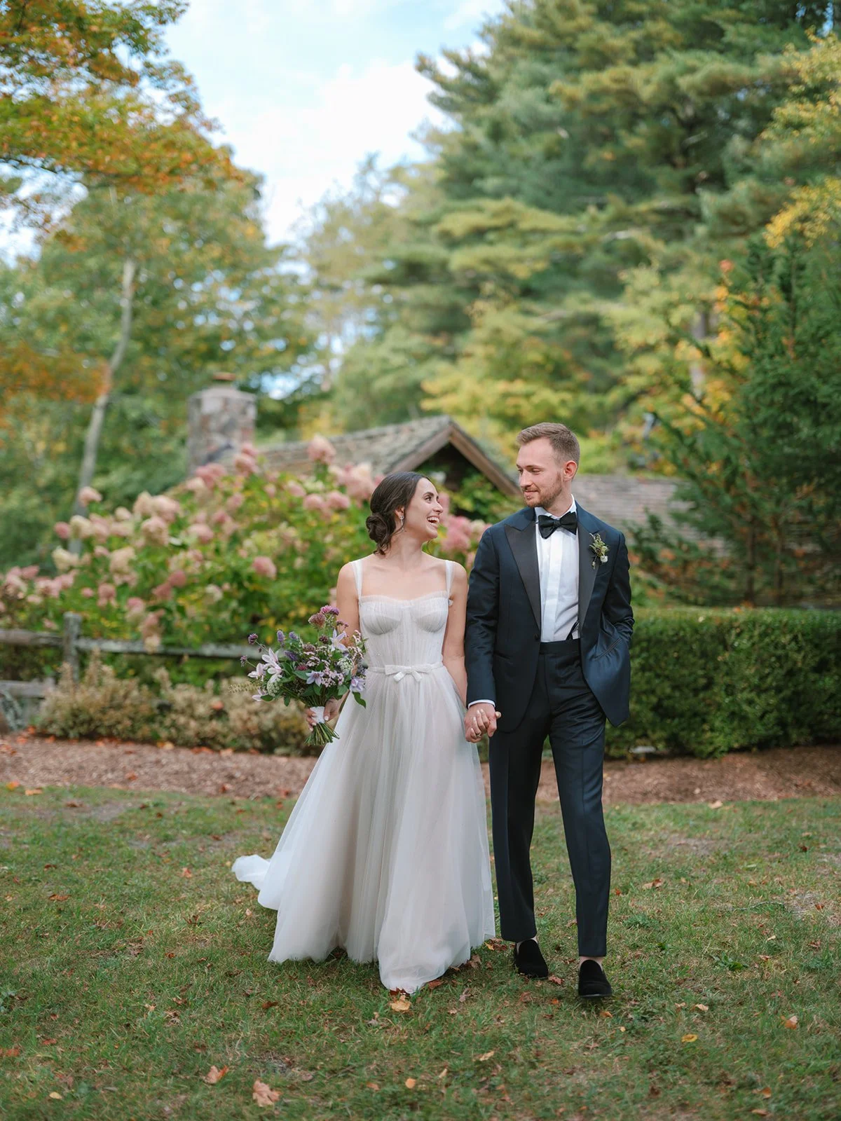 A bride and groom walking hand in hand outdoors, smiling at each other, surrounded by lush greenery and flowering bushes.