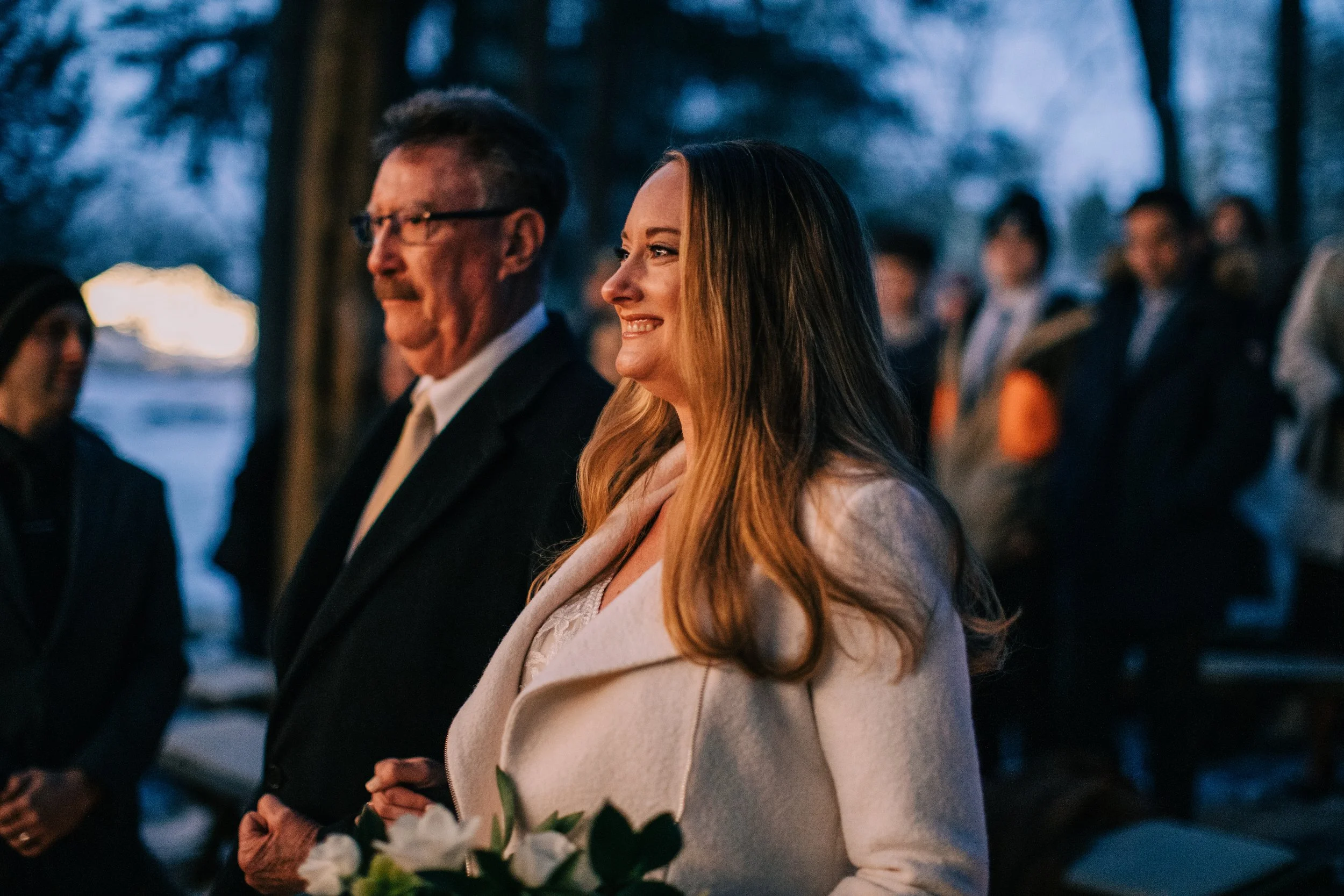 A woman with long hair in a light-colored coat smiling at an outdoor event during dusk, with a man in a dark suit next to her and several people in the background.