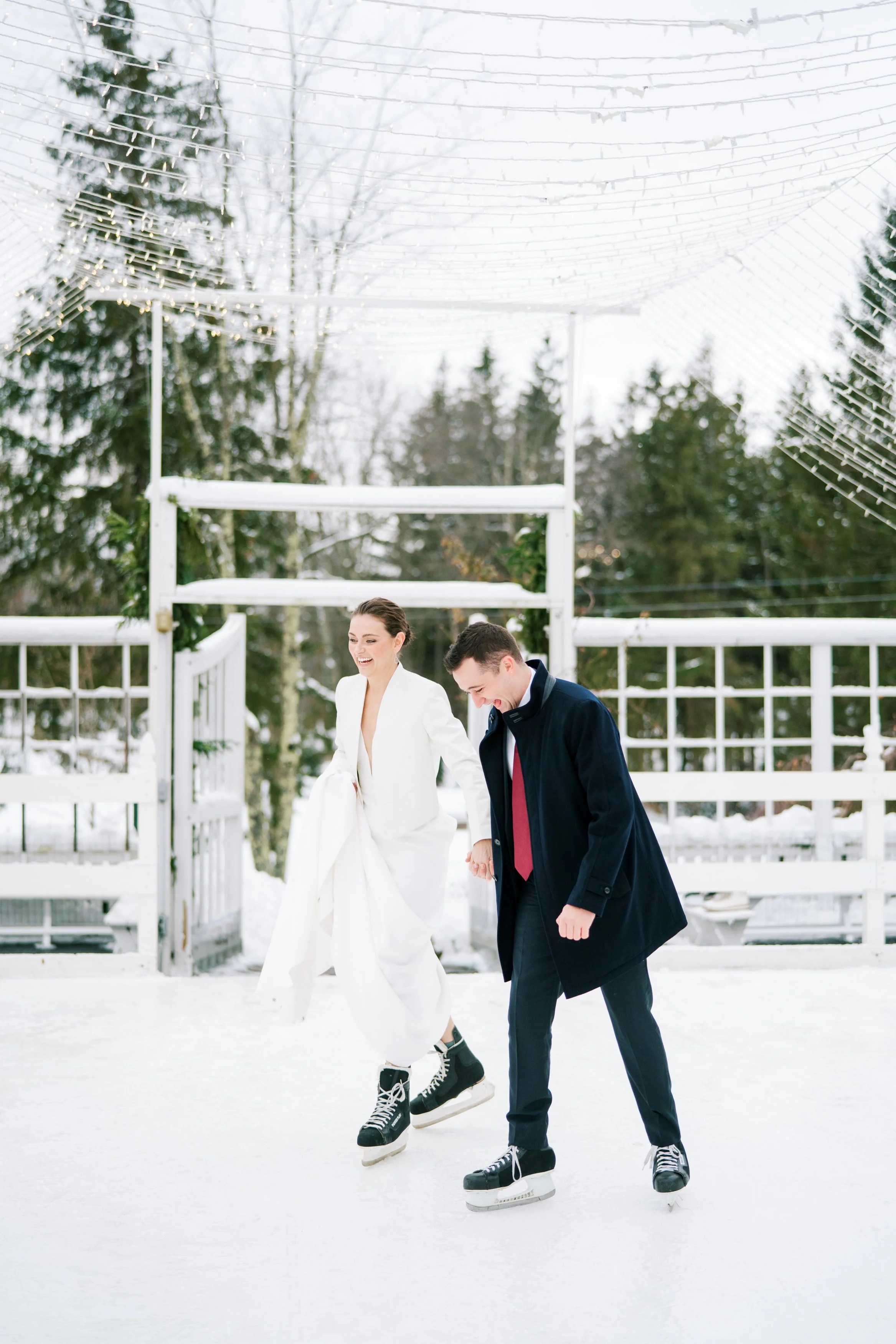 A couple ice skating outdoors in winter, with Christmas lights and snow-covered trees in the background.