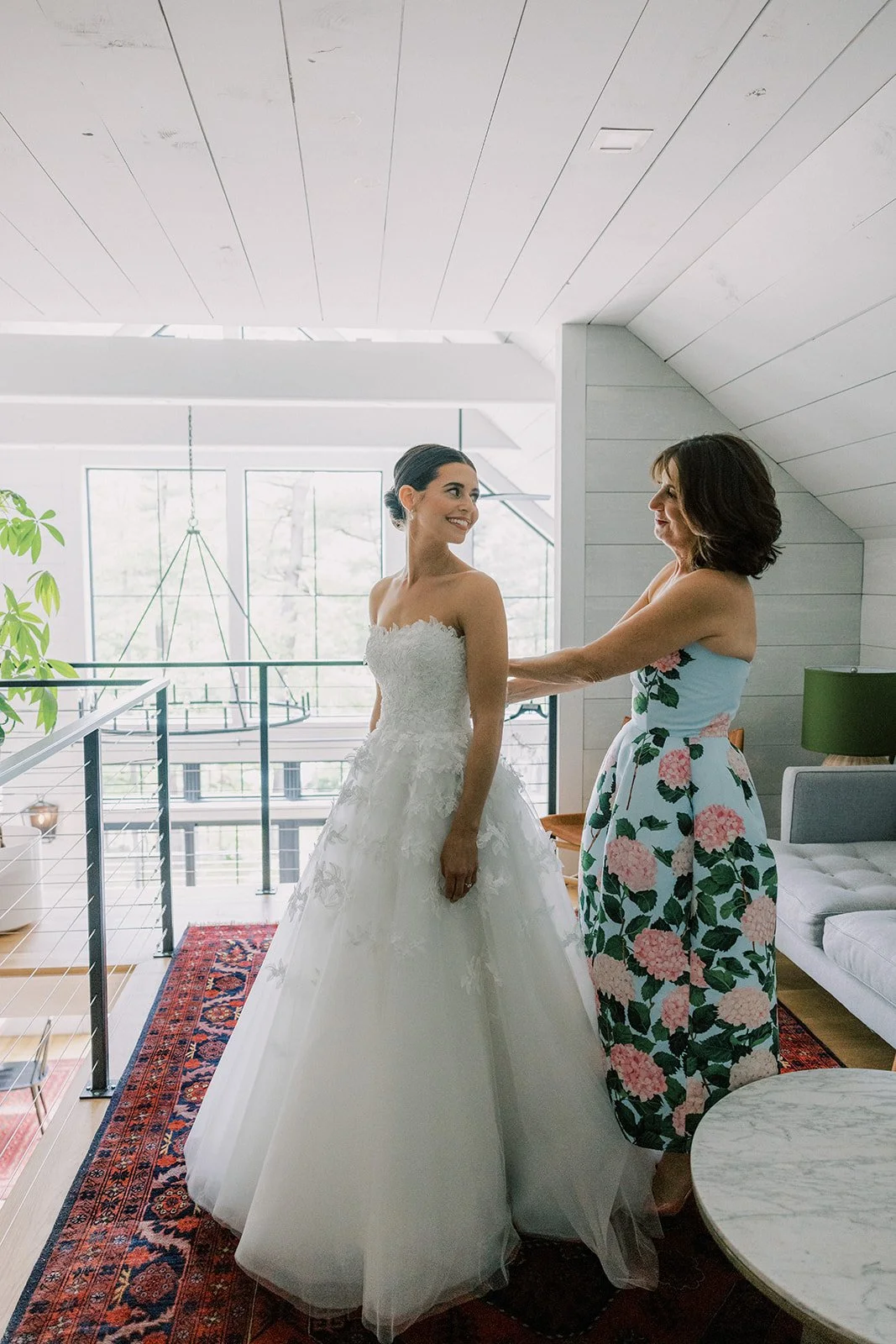 A bride in a white wedding dress is smiling at a woman in a floral dress inside amodern room with large windows and a red patterned rug.
