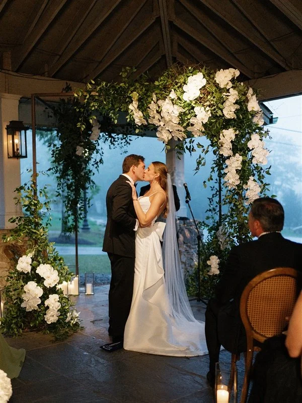 A bride and groom share a kiss during their wedding ceremony under a floral arch with white roses and greenery. An officiant and guests are also visible, seated and observing.