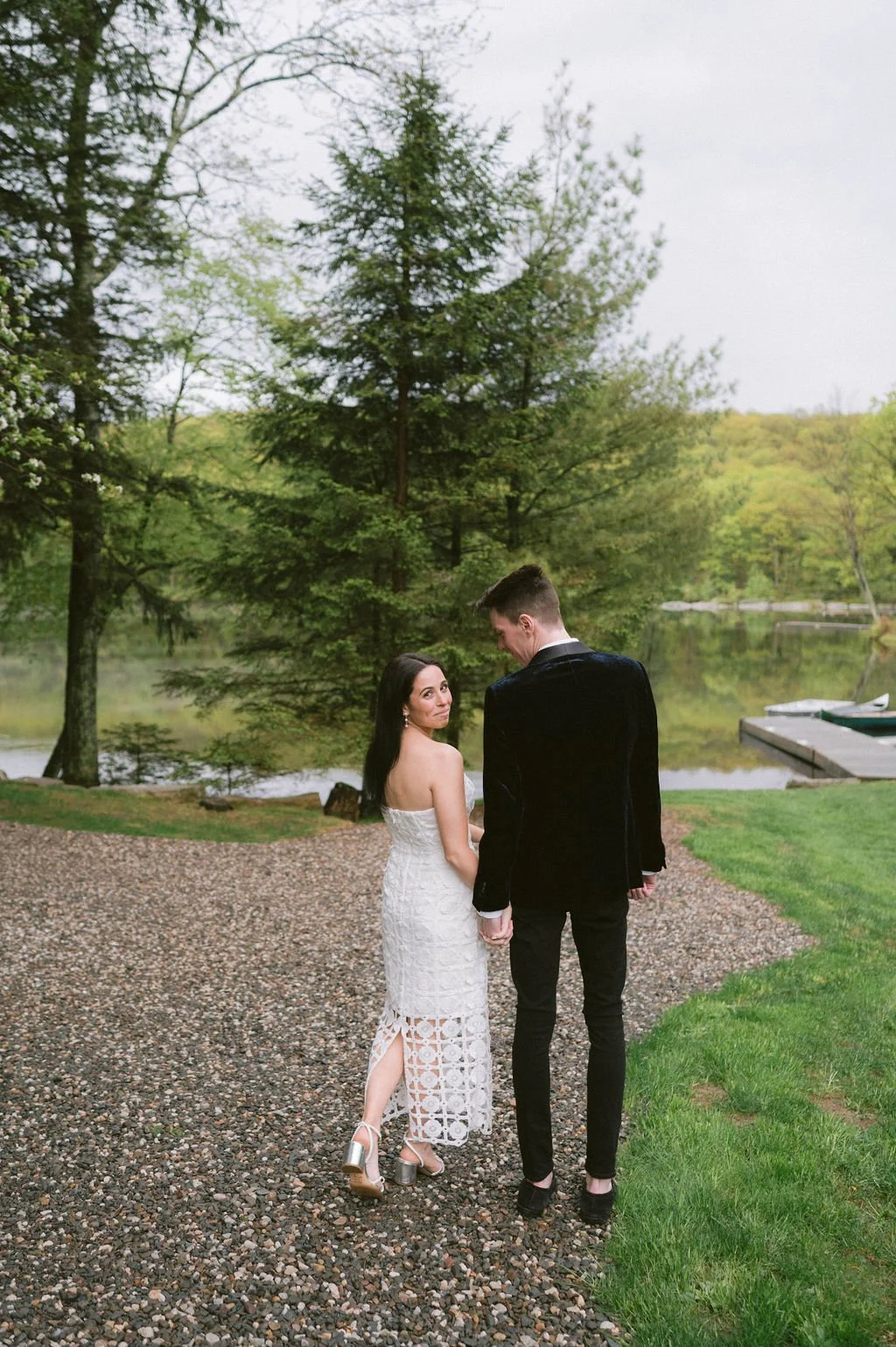 A couple in wedding attire standing on a gravel path near a lake, with trees and green foliage in the background.