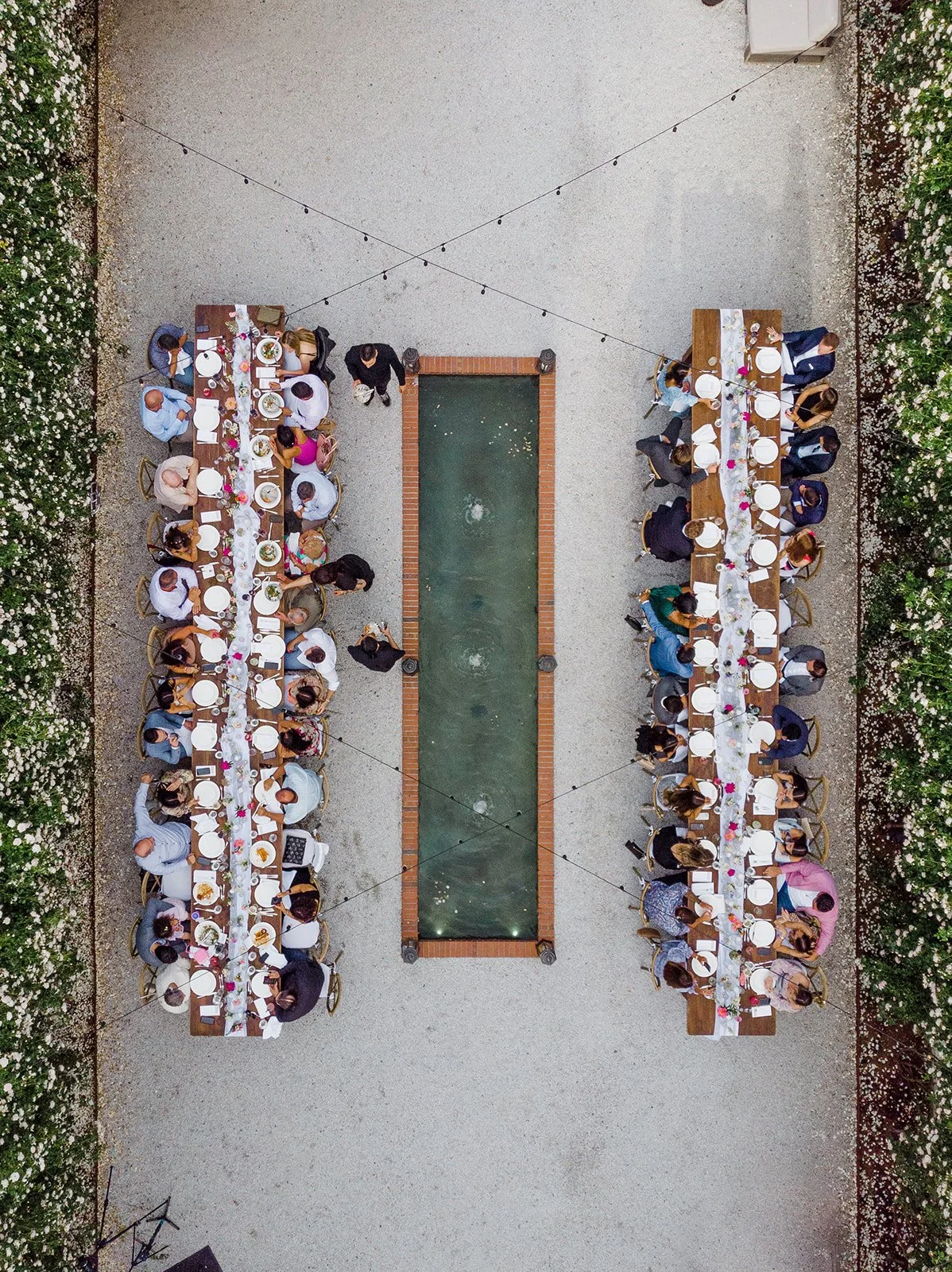 An aerial view of a large outdoor dinner party with two long tables set on either side of a rectangular pond, surrounded by flowering bushes.