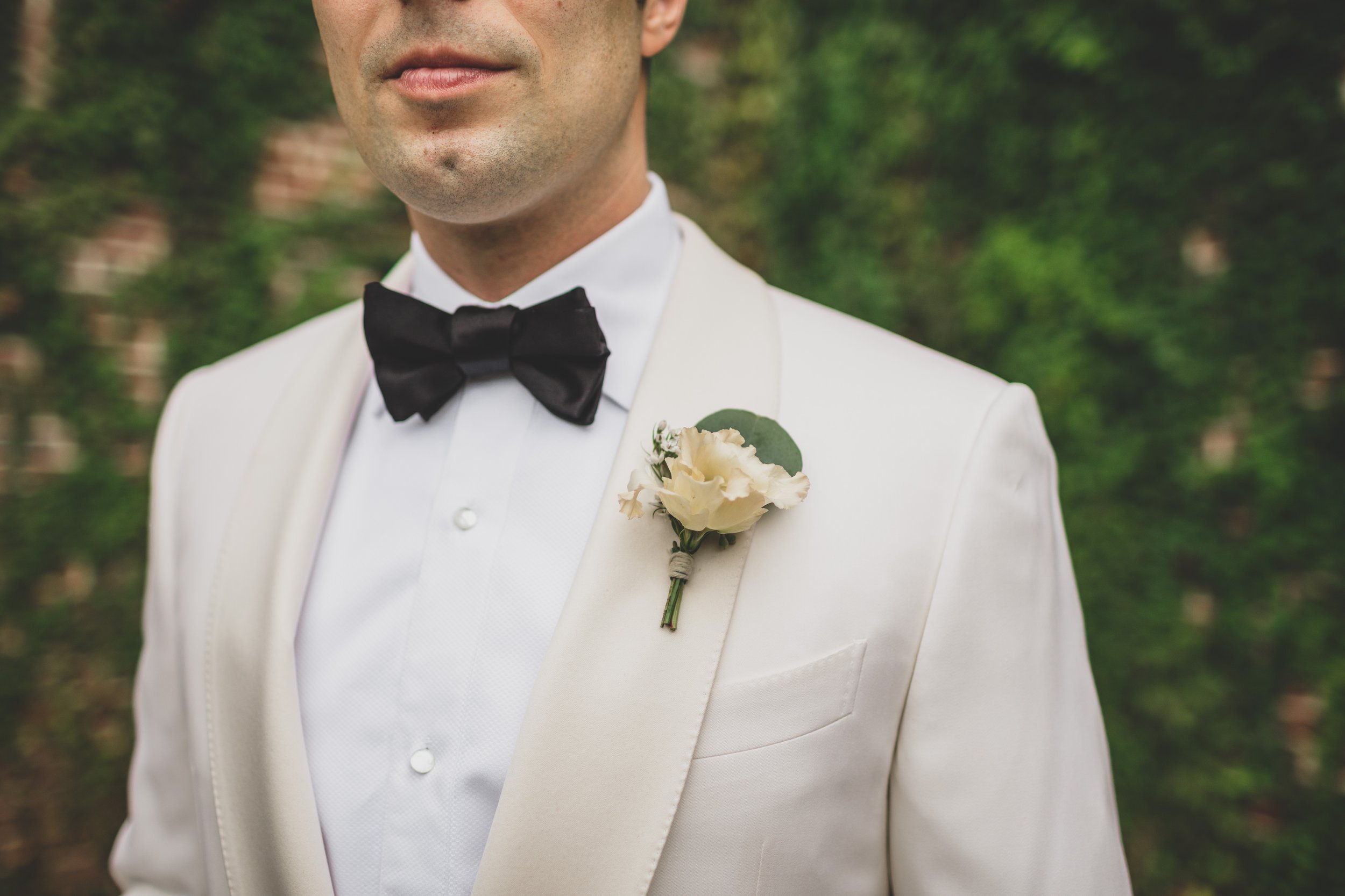 Close-up of a groom in a white tuxedo with a black bow tie and a white boutonniere on his lapel, standing outdoors with greenery in the background.