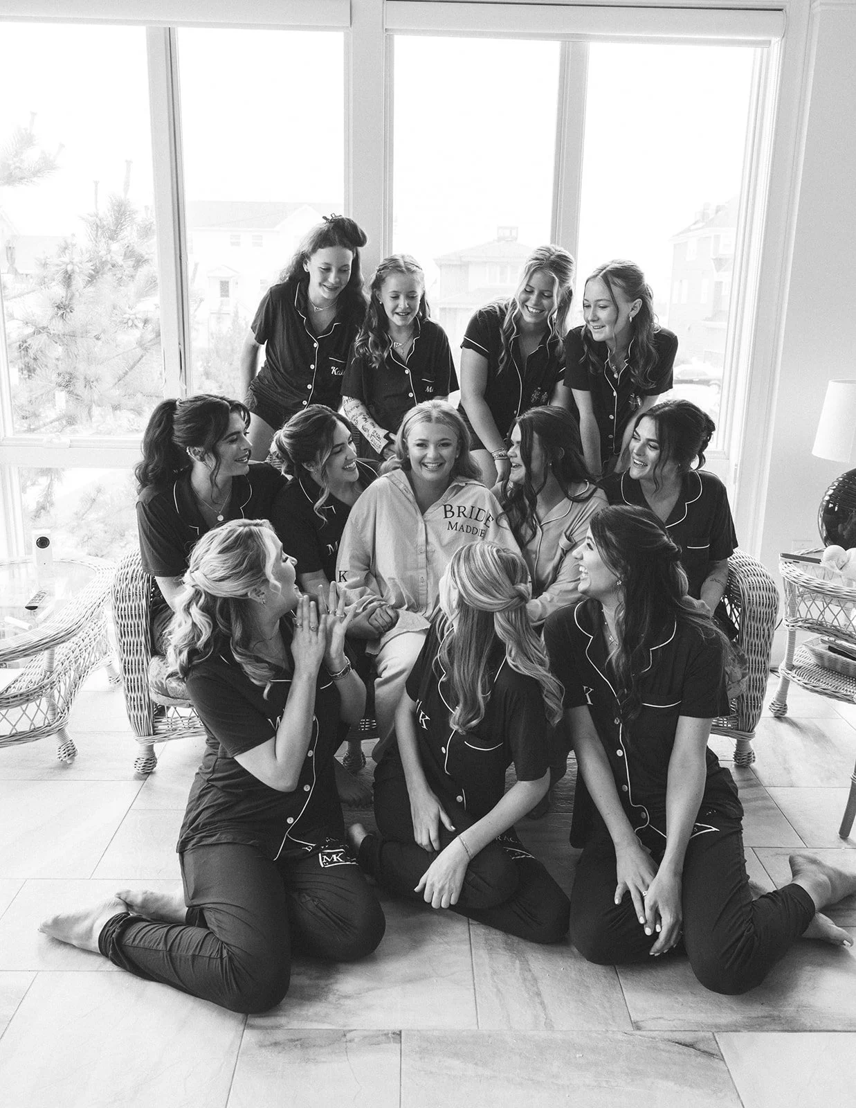 A group of women, including a bride, gathering for a pre-wedding ritual in a bright room with large windows, wearing matching pajamas, with some kneeling and others standing around the bride.