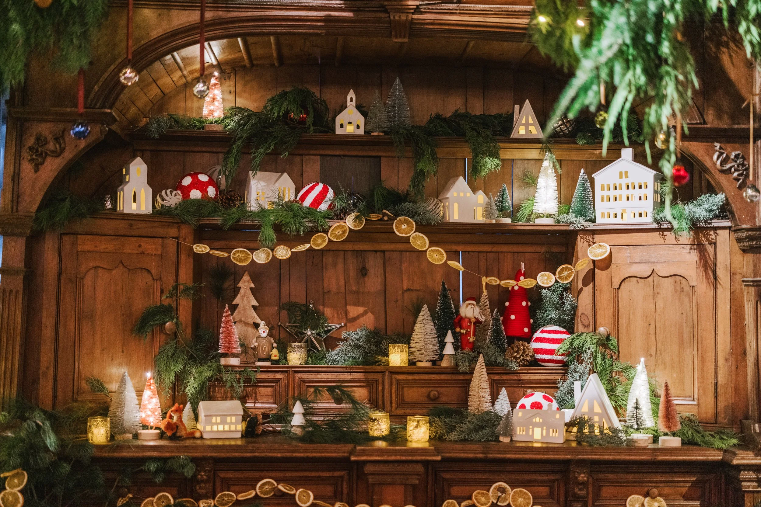 Christmas decoration display with miniature paper houses, ornaments, pine branches, and small decorative trees on wooden shelves.