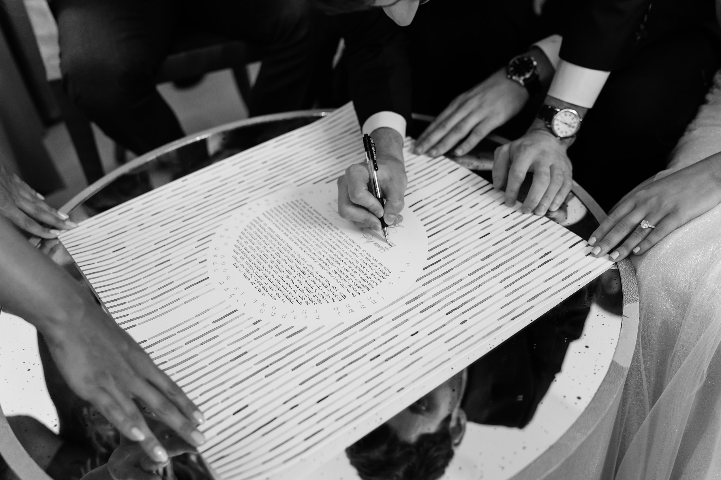 People gather around a table as one person signs a document, with visible hands and watches, in a formal setting.