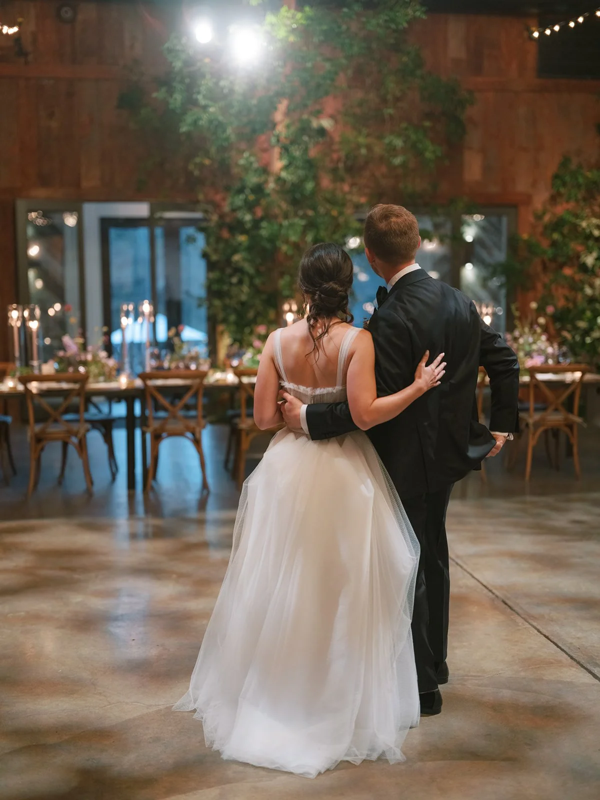 A bride and groom are dancing together in a wedding reception hall, with a decorated table in the background and a wall covered with greenery and flowers.