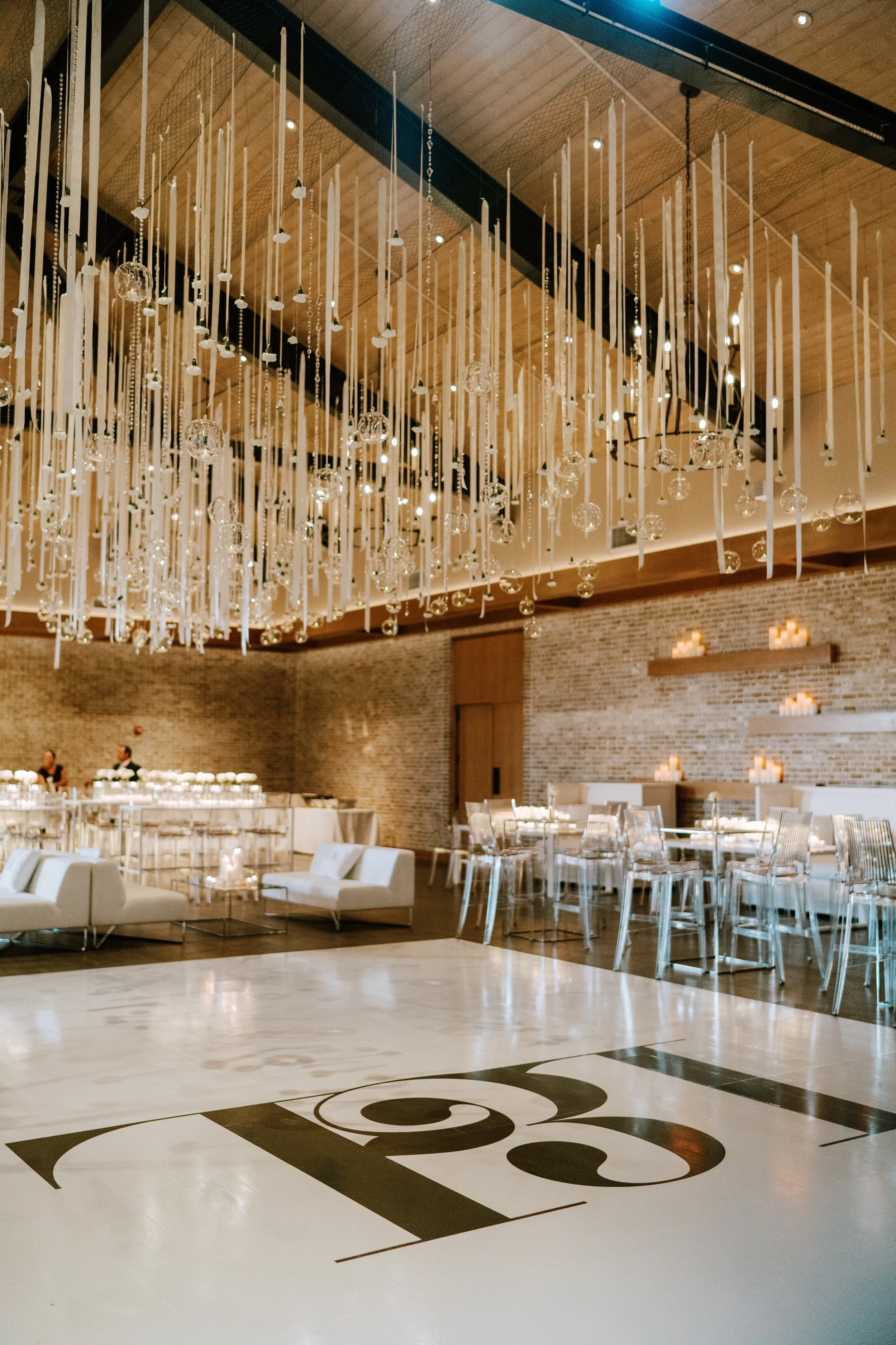 Elegant event space with hanging chandeliers, white seating, glass chairs, and candlelit walls.