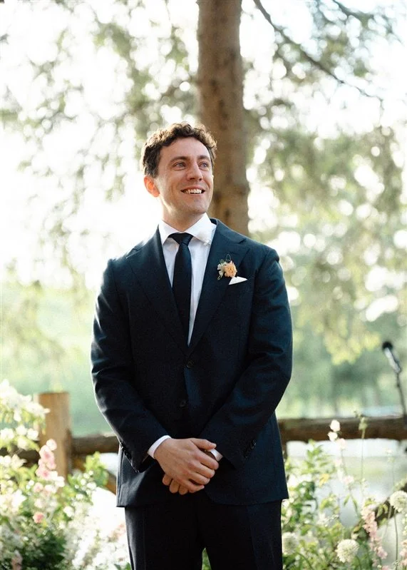 A smiling man in a dark suit and tie standing outdoors with trees and flowers in the background.