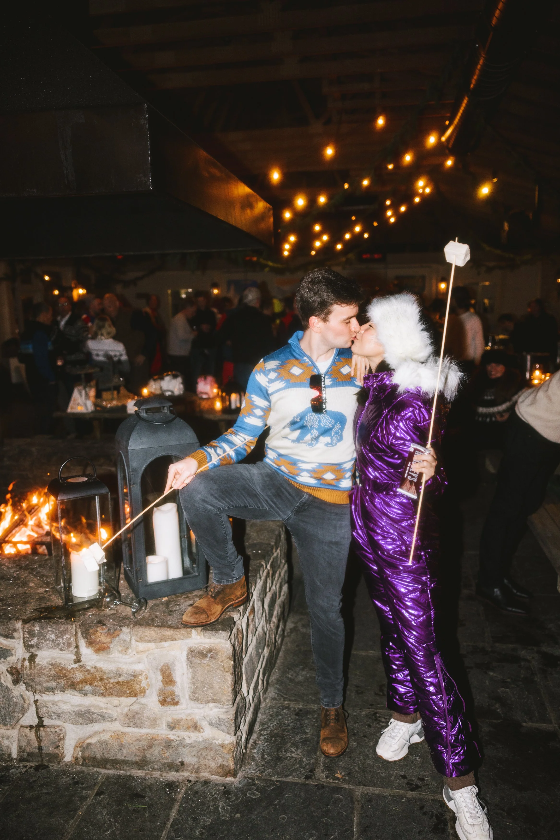 A young couple sharing a kiss at a festive indoor holiday party, with string lights and a fireplace in the background, surrounded by other guests.