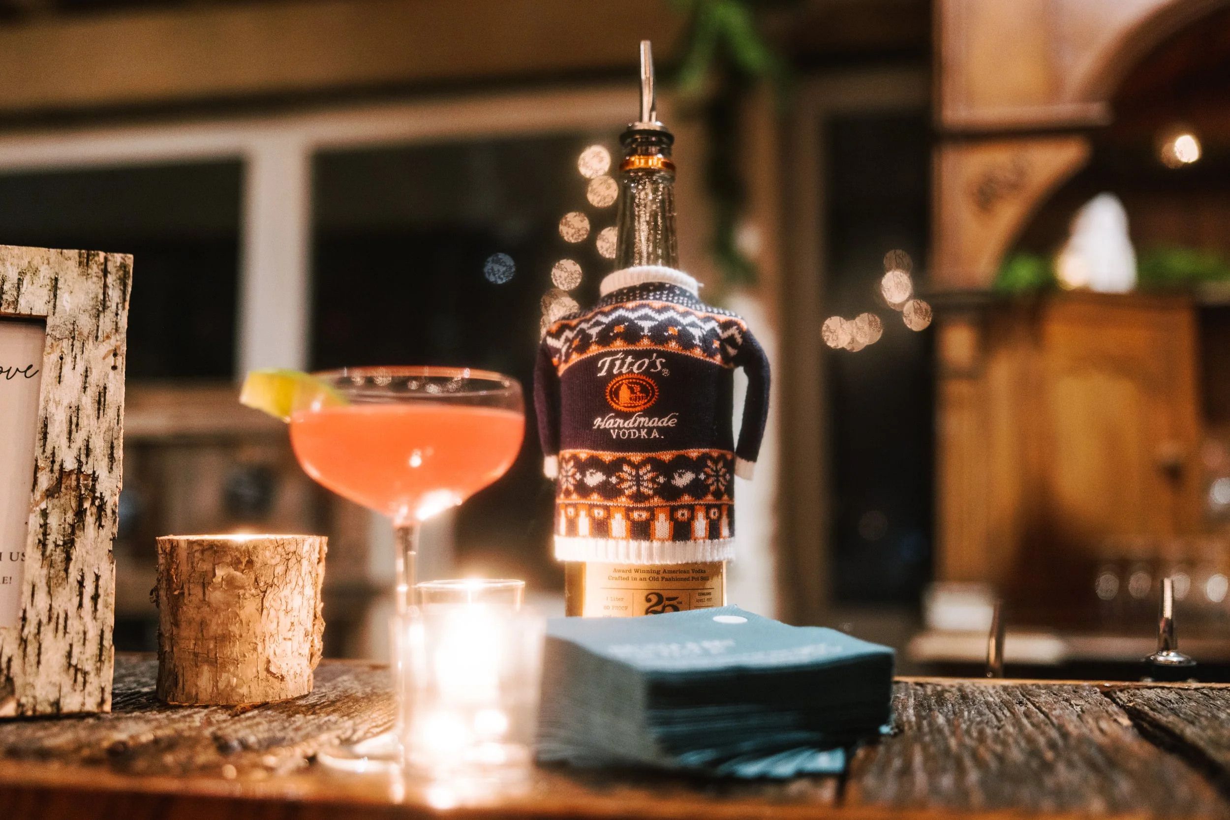 A pink cocktail with a lemon wedge on a wooden table, a bottle of Tito's handmade vodka with a festive sweater cap, a small candle, and a stack of napkins, in a cozy, warmly-lit bar or restaurant setting.