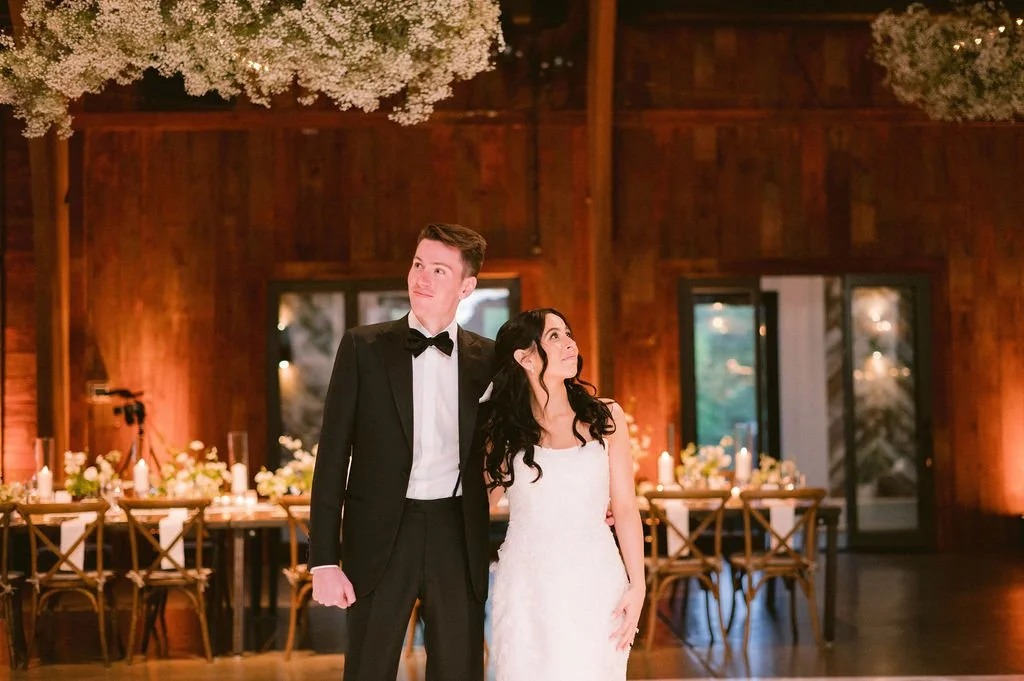 A bride and groom standing together at a wedding reception in a rustic wooden venue, with floral decorations and candles on a table in the background.