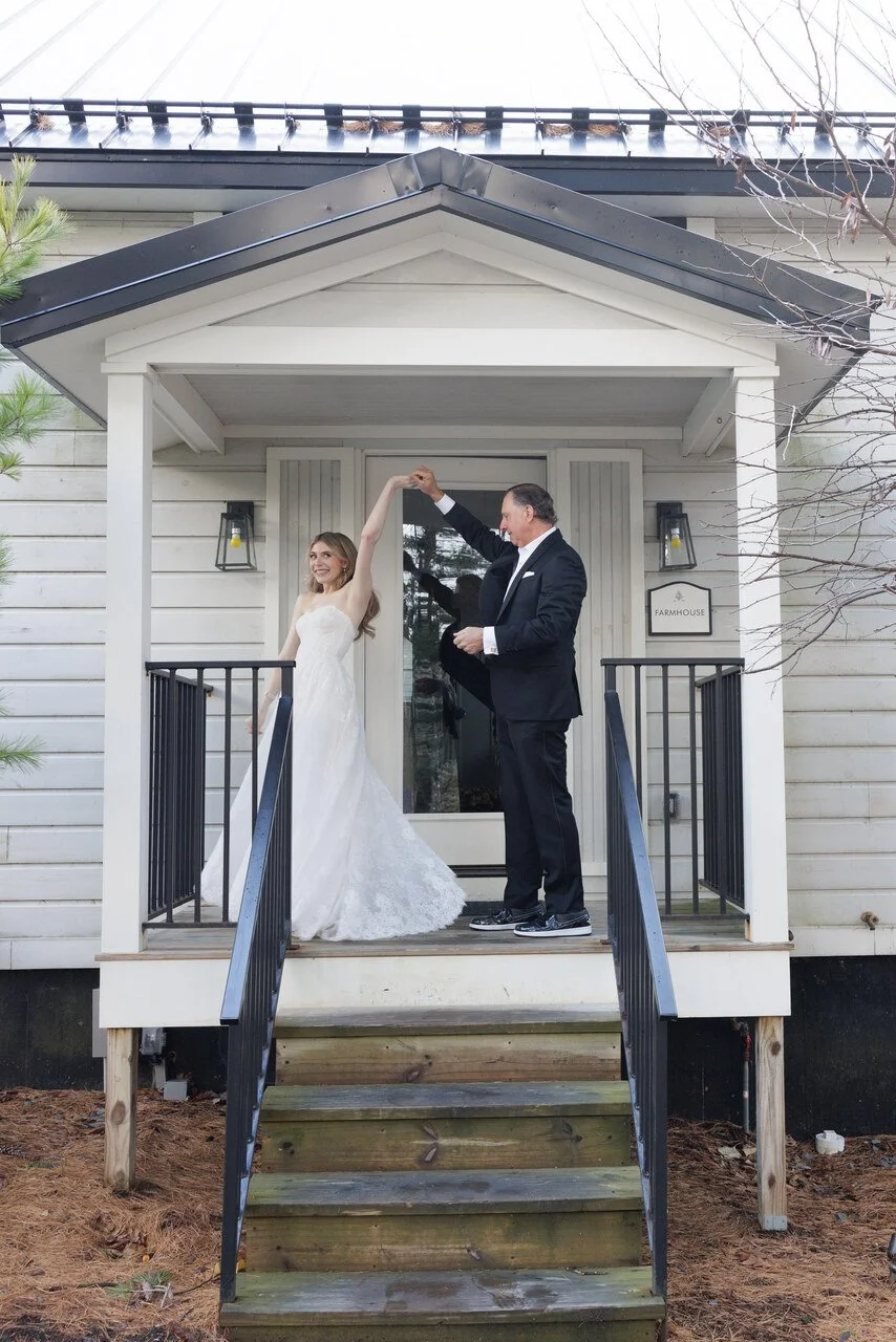 A bride and groom dancing on the front porch of a farmhouse, with the groom twirling the bride. The porch has black railing, white siding, and a sign that reads 'FARMHOUSE'. The bride is in a strapless white wedding dress, and the groom is in a black