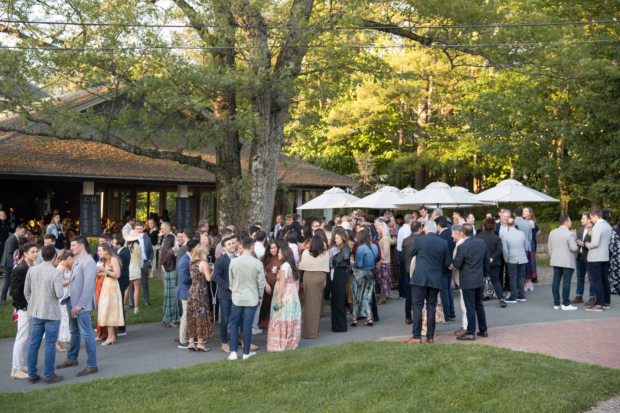 Guests mingling at an outdoor social event or wedding reception under large white umbrellas, with trees and a building in the background.