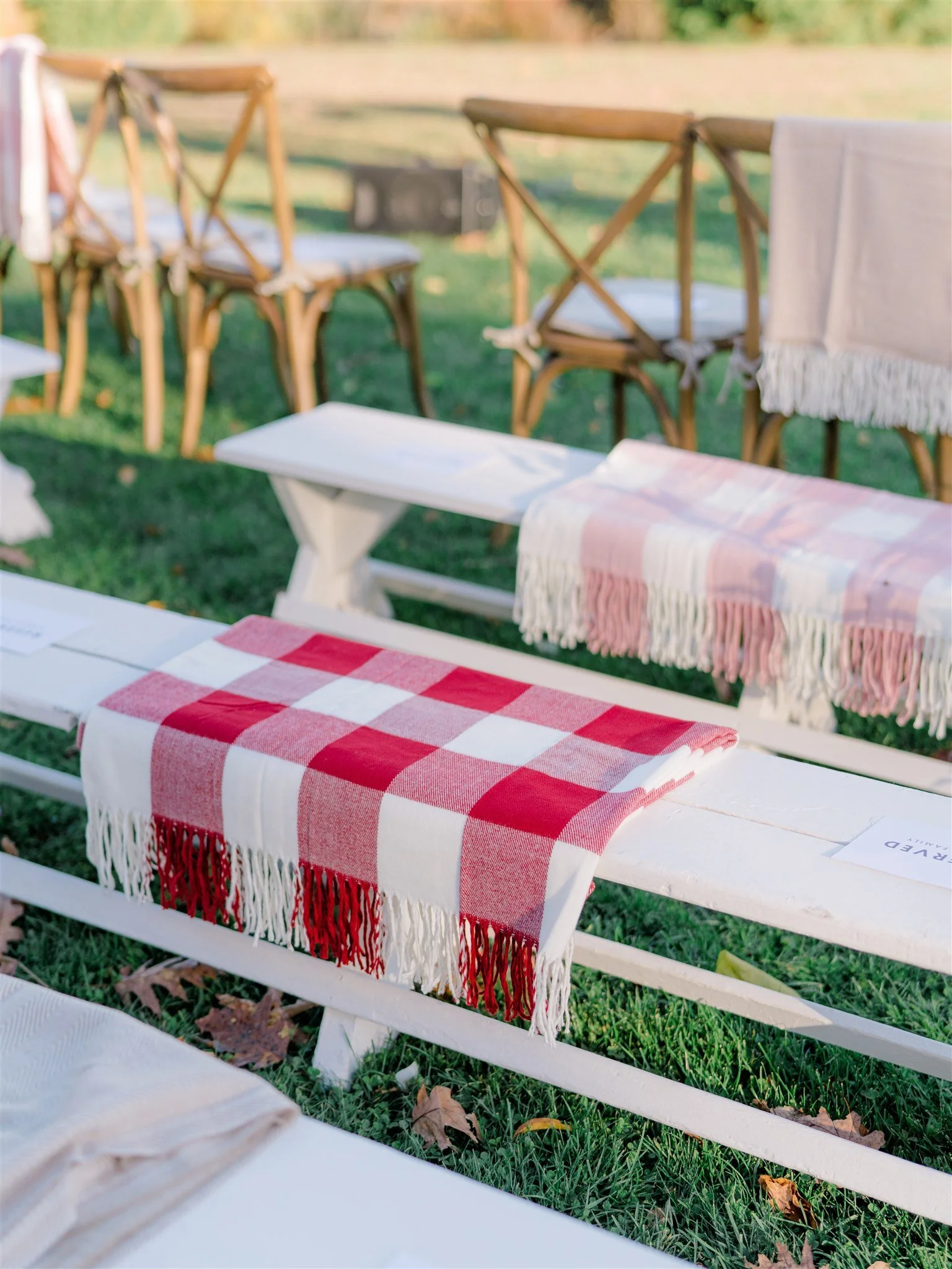 Outdoor event setup with white benches, red and white checkered tablecloths, and wooden chairs on grass.