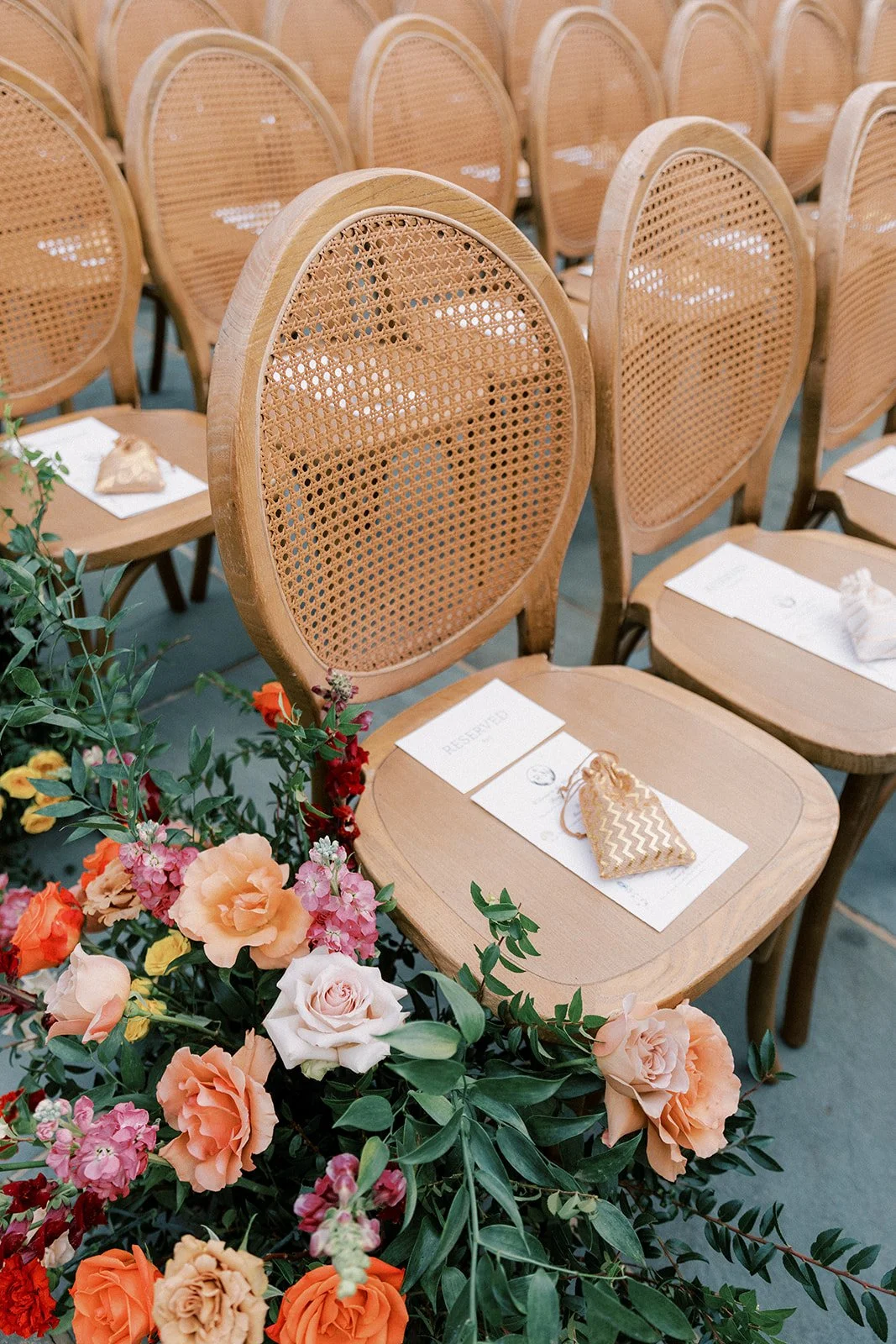 Set of wooden chairs with woven backrests arranged in a row, reserved sign on one chair, and floral arrangement with pink, orange, and white roses in the foreground.