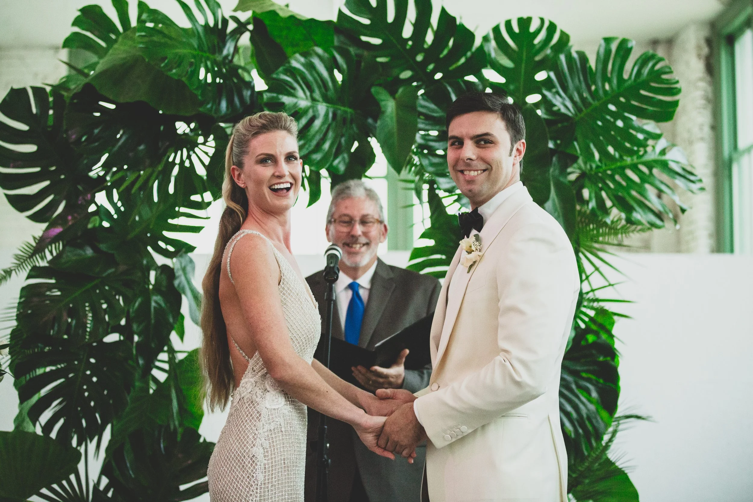A wedding ceremony with a bride and groom holding hands and smiling at each other indoors, with a person officiating and a green leafy plant background.