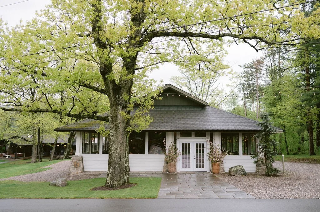 A house with a dark roof, white exterior, surrounded by trees, with a stone pathway and potted plants at the entrance