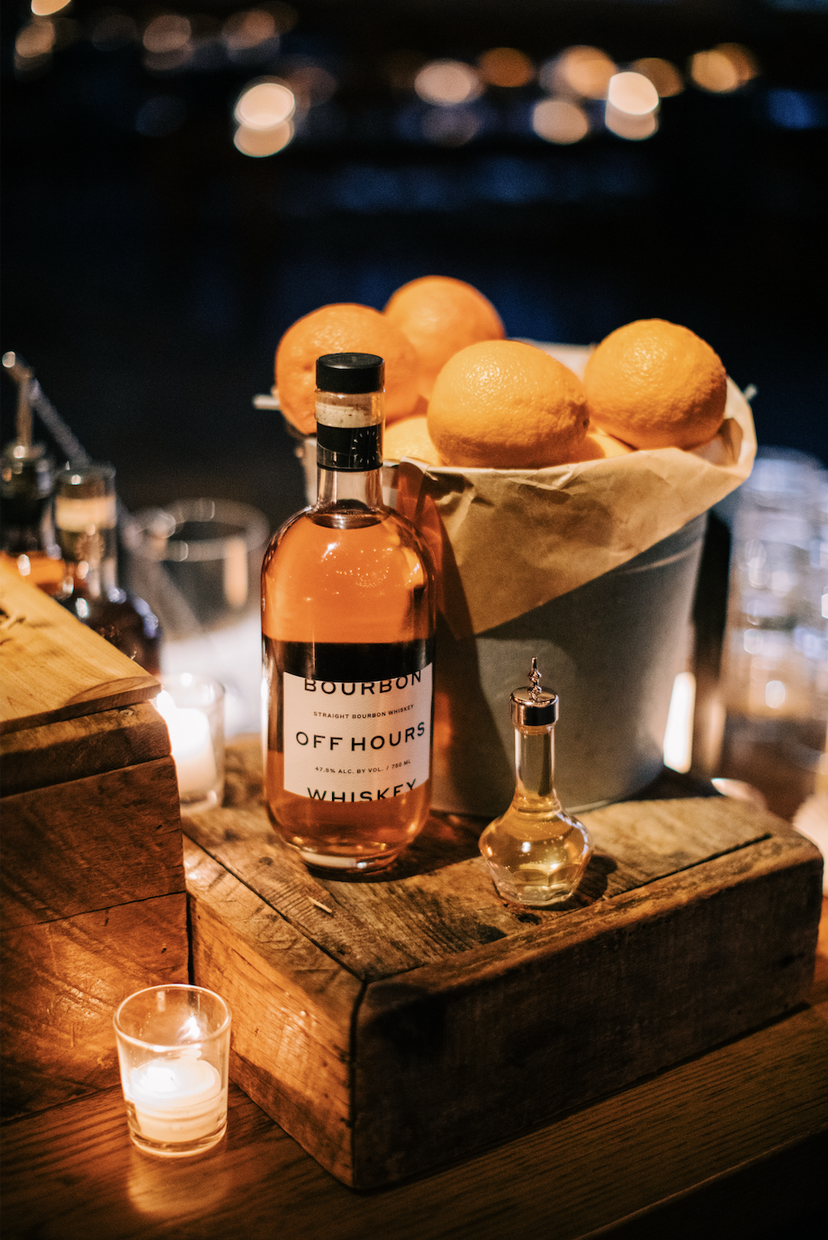 Bottle of bourbon whiskey, a basket of lemons, and small glass bottles on a rustic wooden bar counter at night with blurred city lights in the background.