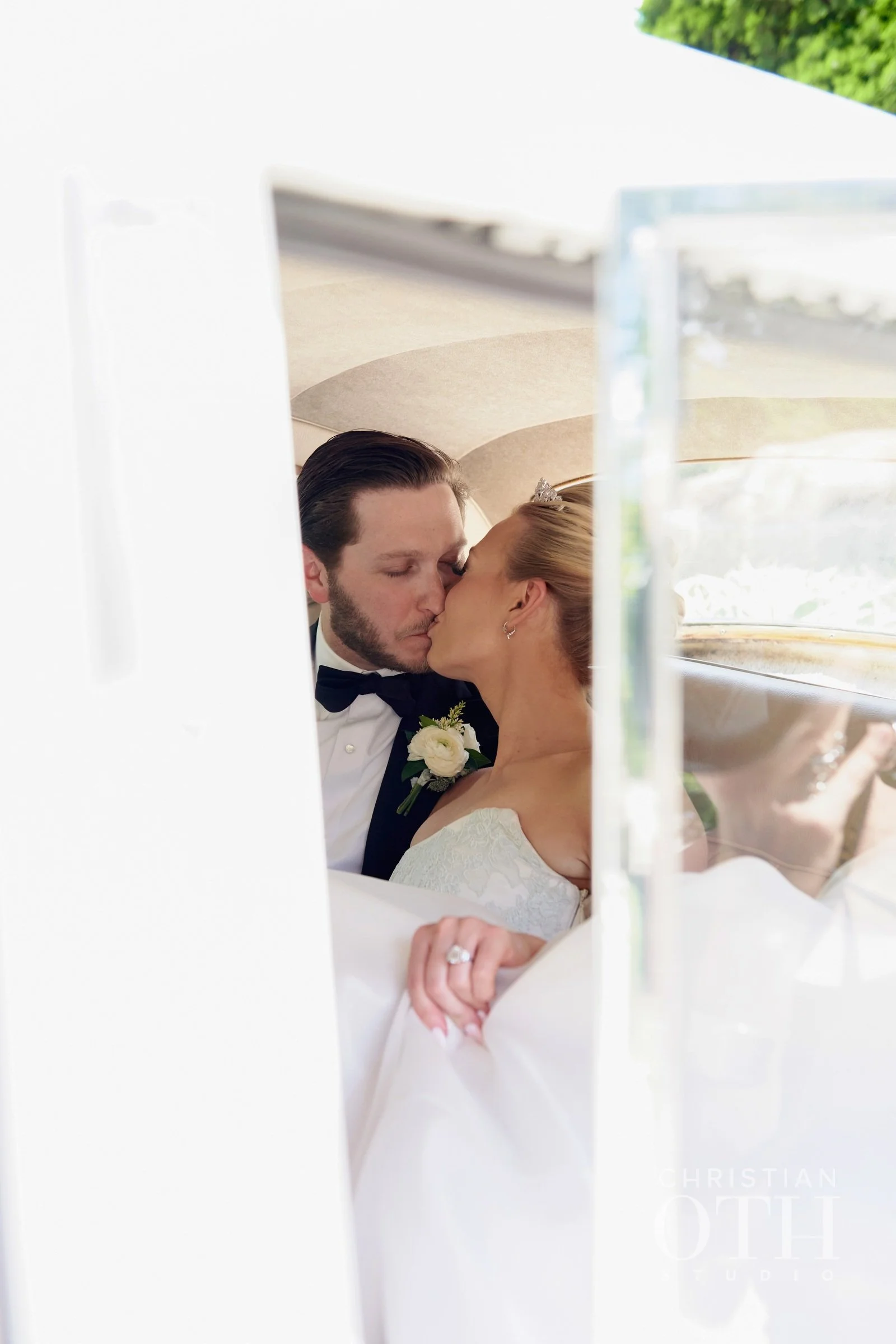 A bride and groom sharing a first kiss, seen through the window of a vehicle, dressed in wedding attire.