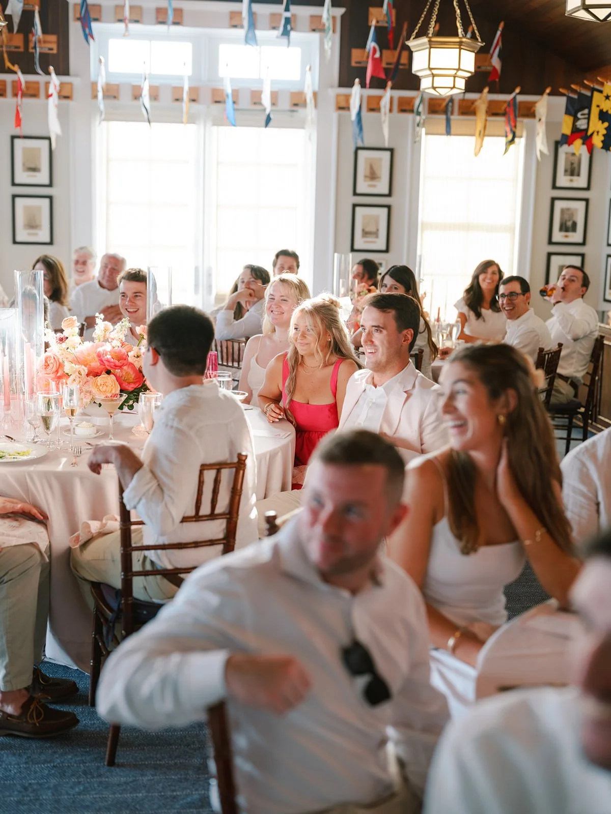 Guests sitting at a long dining table during a celebration or wedding reception, with sunlight streaming through large windows, colorful flags hanging from the ceiling, and a floral centerpiece on the table.