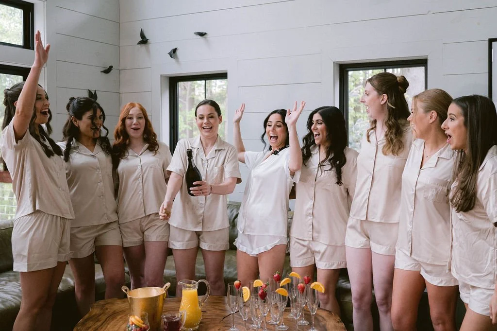 A group of women in pajamas celebrating, some with raised hands, holding drinks, near a table with pitchers and glasses, in a bright room with windows and wall decorations.