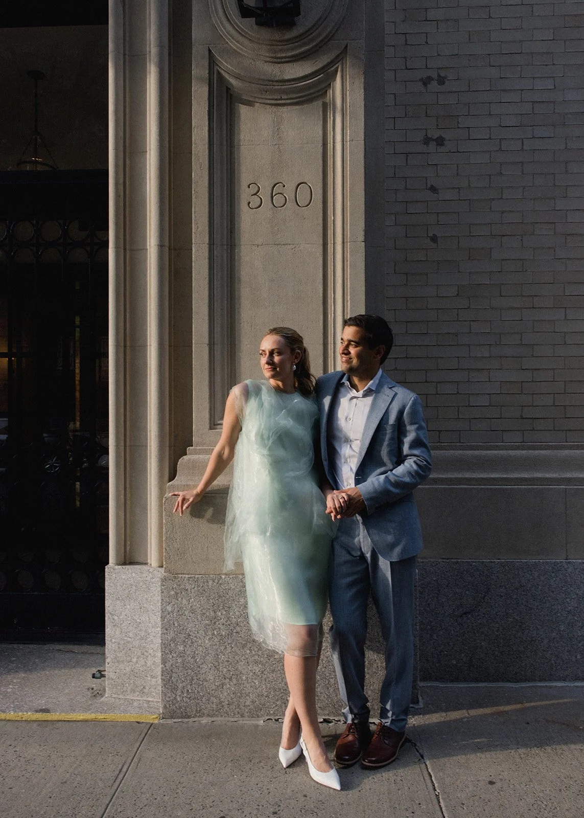 A couple in formal attire standing outside a building with the address 360 inscribed on it. The woman is wearing a light-colored dress and white high heels, leaning against the wall. The man is in a blue suit with a white shirt, standing close to her