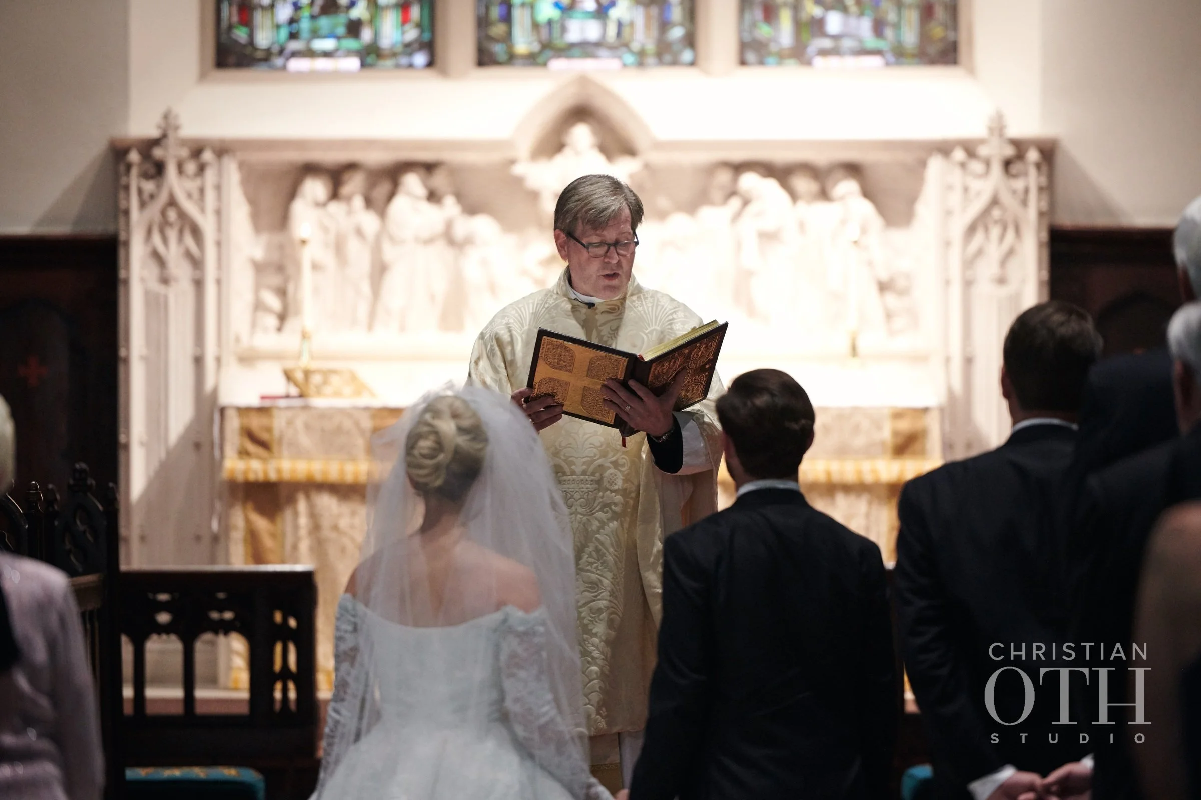 A priest reads from a book during a wedding ceremony in a church, with a bride in a veil and wedding gown and groom standing before him, and guests seated in pews.