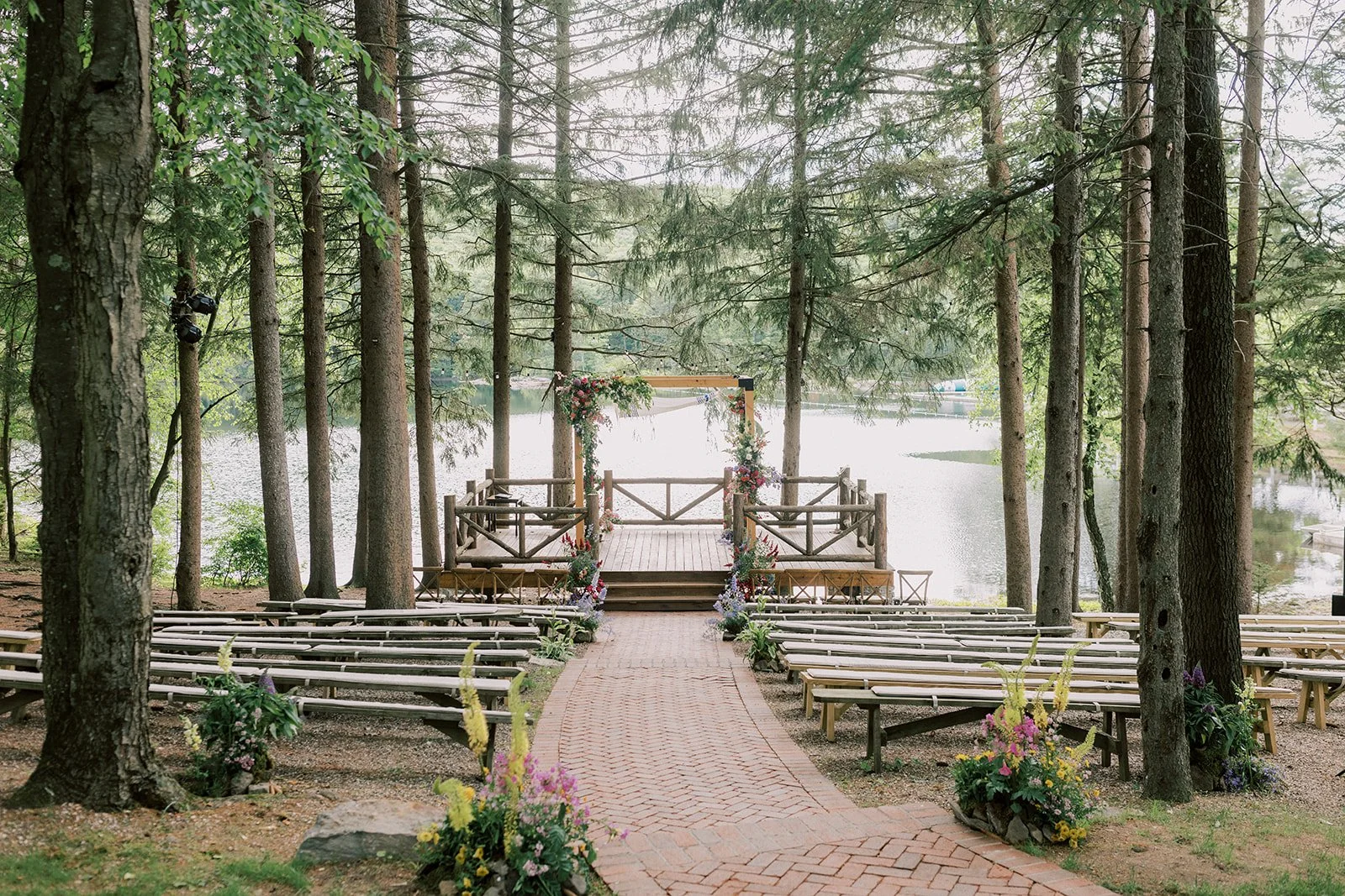 A wedding ceremony setup outdoors by a lake, with wooden benches on either side of a brick pathway leading to a decorated wedding arch with flowers, surrounded by tall pine trees.