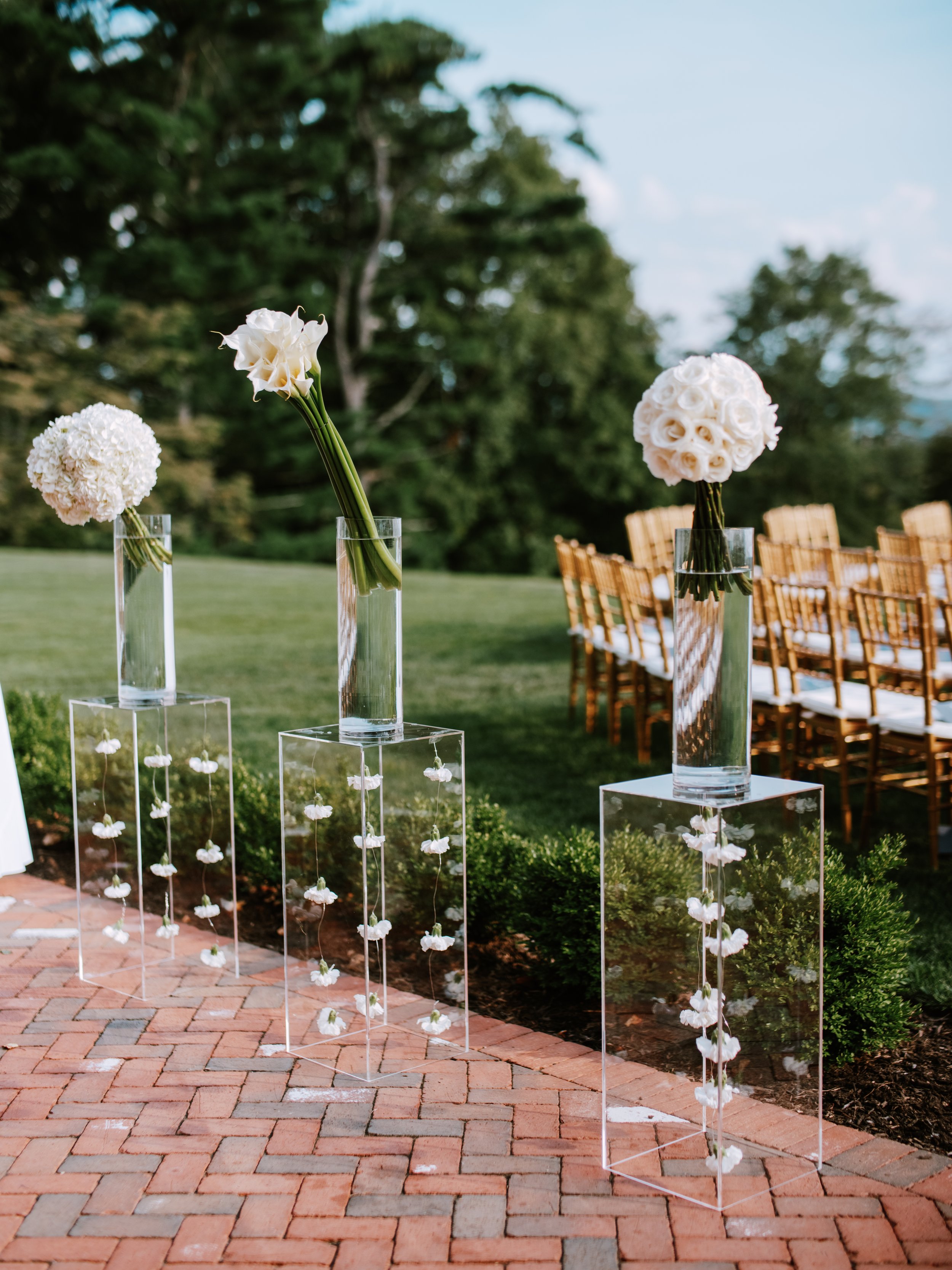 Elegant outdoor wedding setup with three tall glass vases on clear acrylic stands, each holding white floral arrangements, positioned on a brick pathway with green bushes and outdoor chairs in the background.