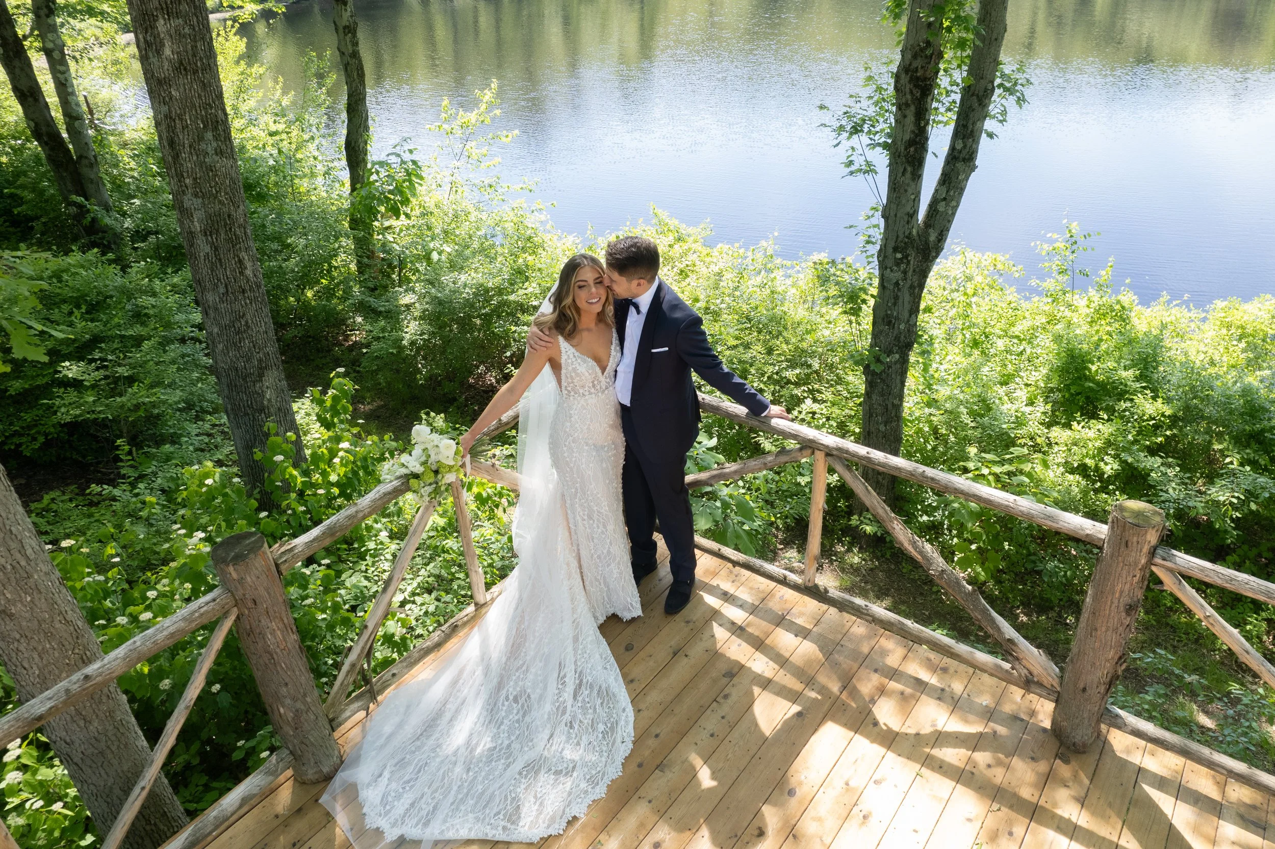 A bride and groom are standing on a wooden deck by a river, surrounded by lush green trees. The bride is wearing a white lace wedding gown, holding a bouquet, and the groom is in a navy suit. The groom is kissing the bride on the forehead as they smi