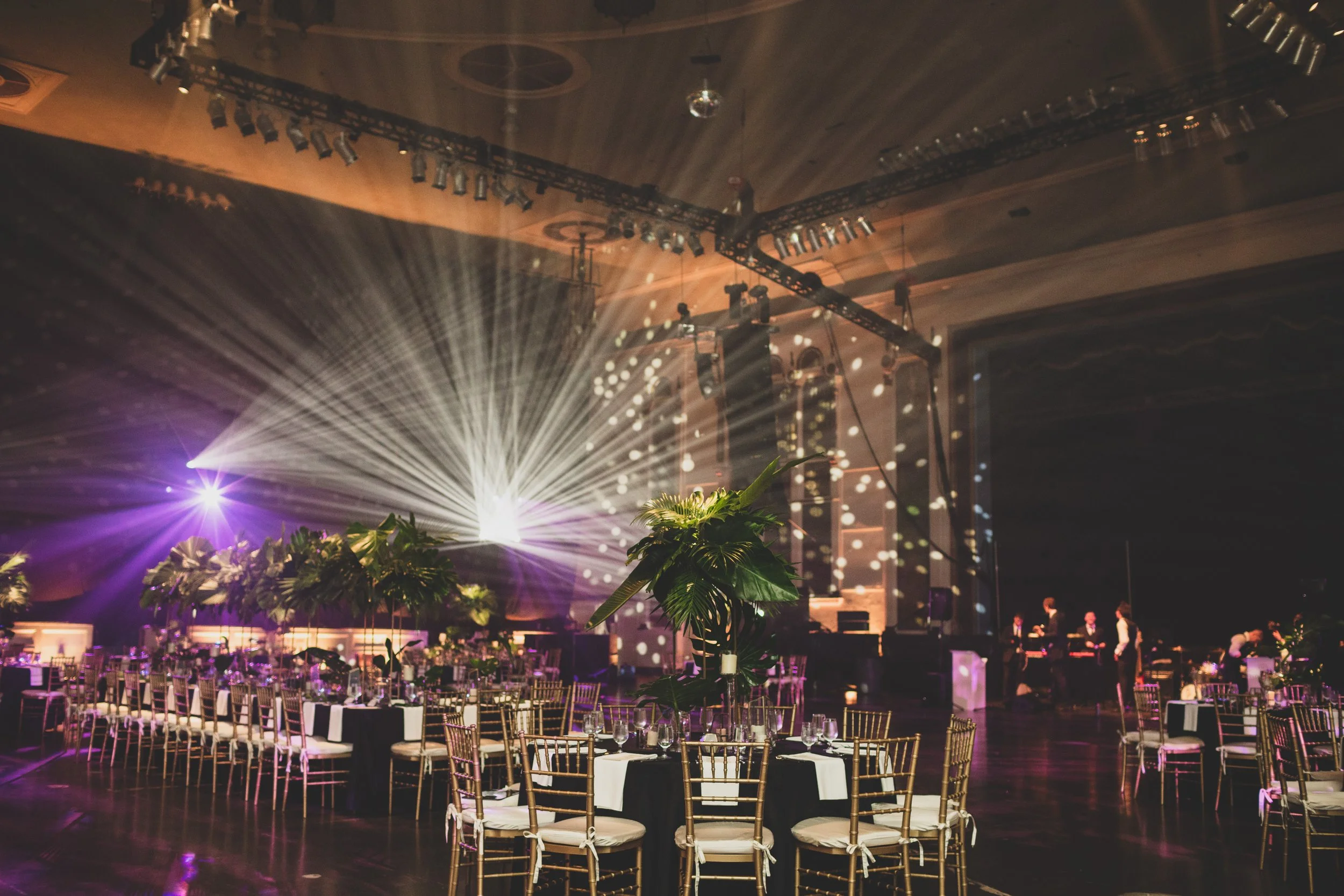 A large event hall with decorated tables and chairs, stage with bright lights, and spectators preparing for a performance or celebration.