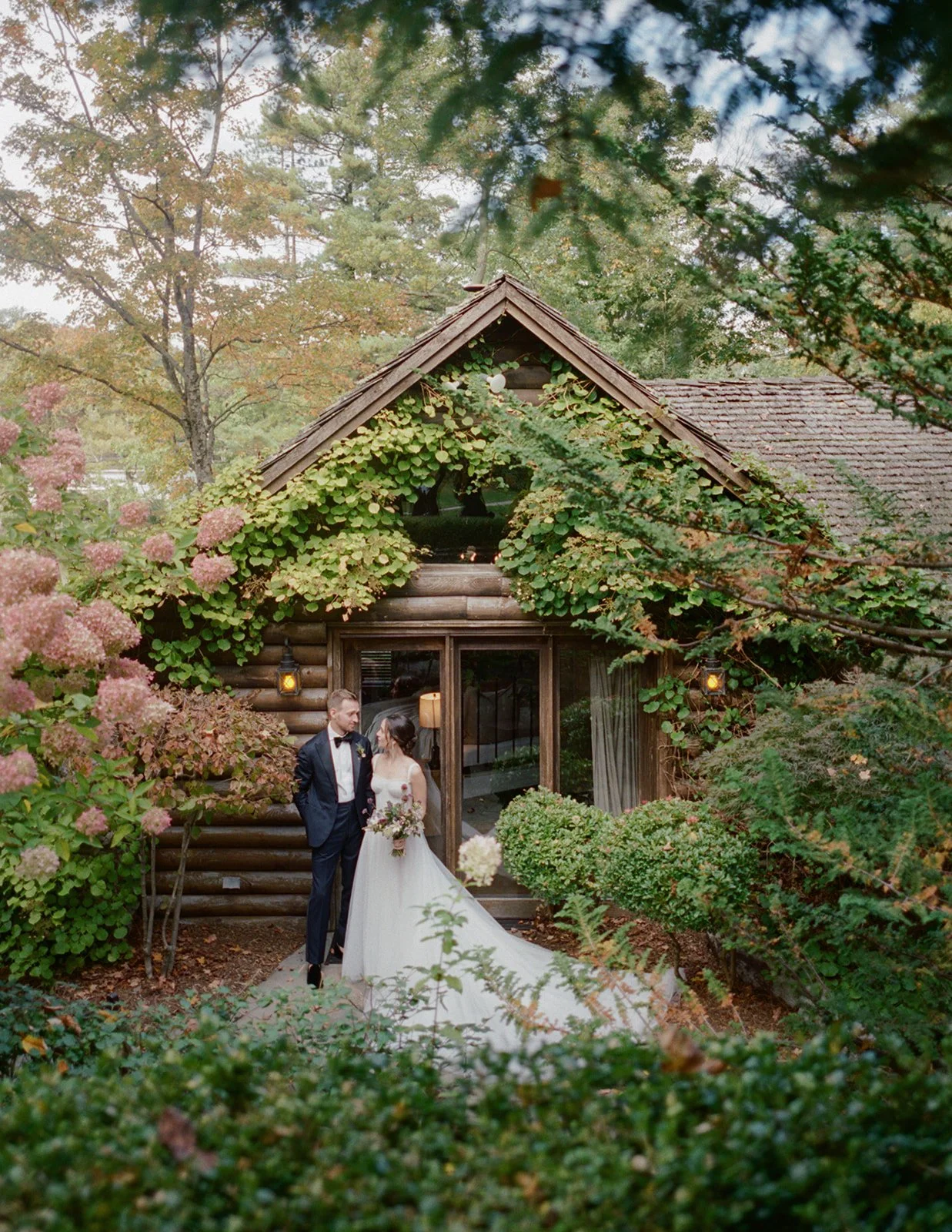 A couple in wedding attire standing outside a rustic log cabin surrounded by lush greenery and pink hydrangeas.