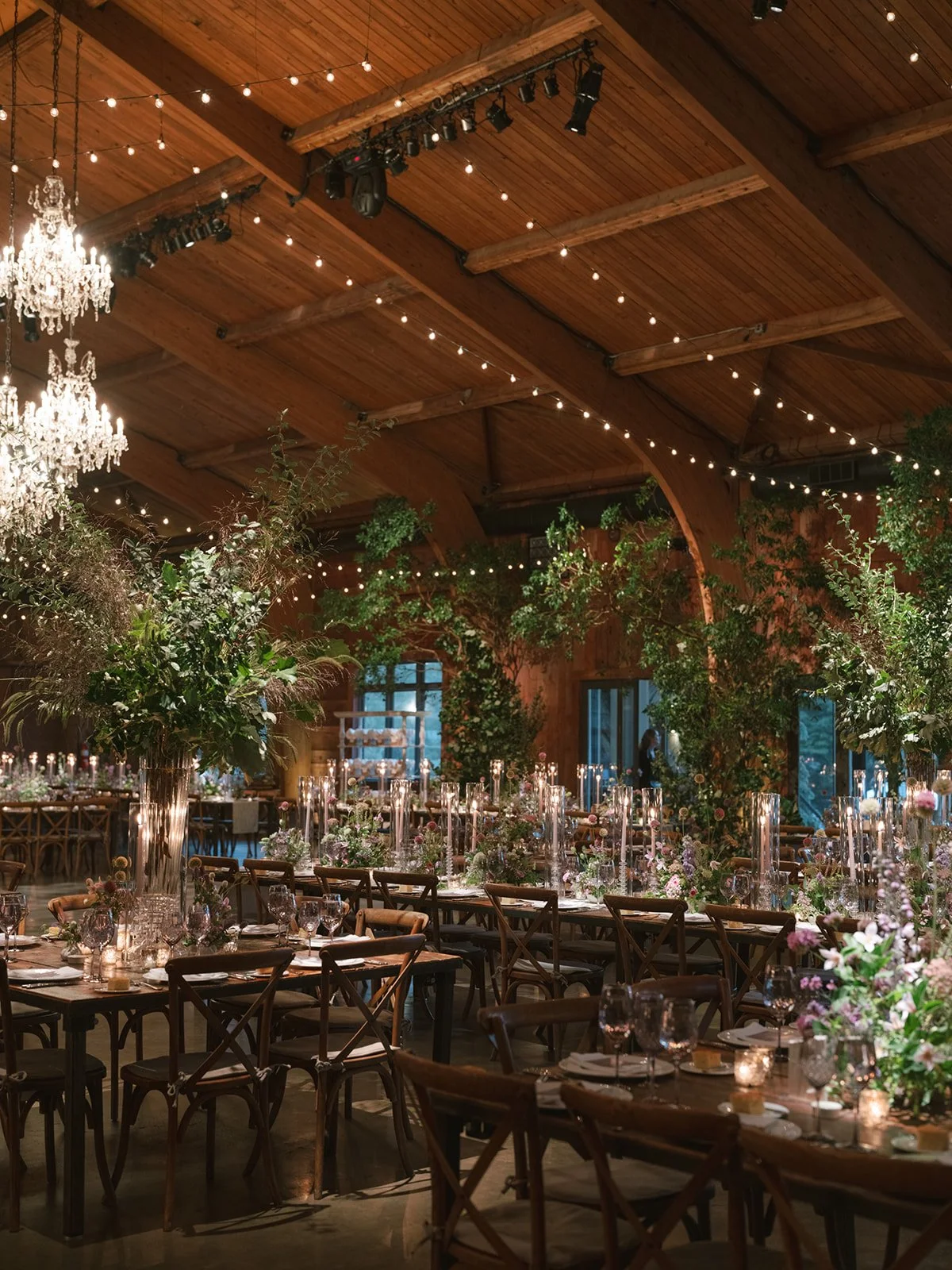 Elegant wedding reception with long tables decorated with candles and floral arrangements, set in a rustic venue with wooden walls, vaulted ceiling, string lights, and chandeliers.