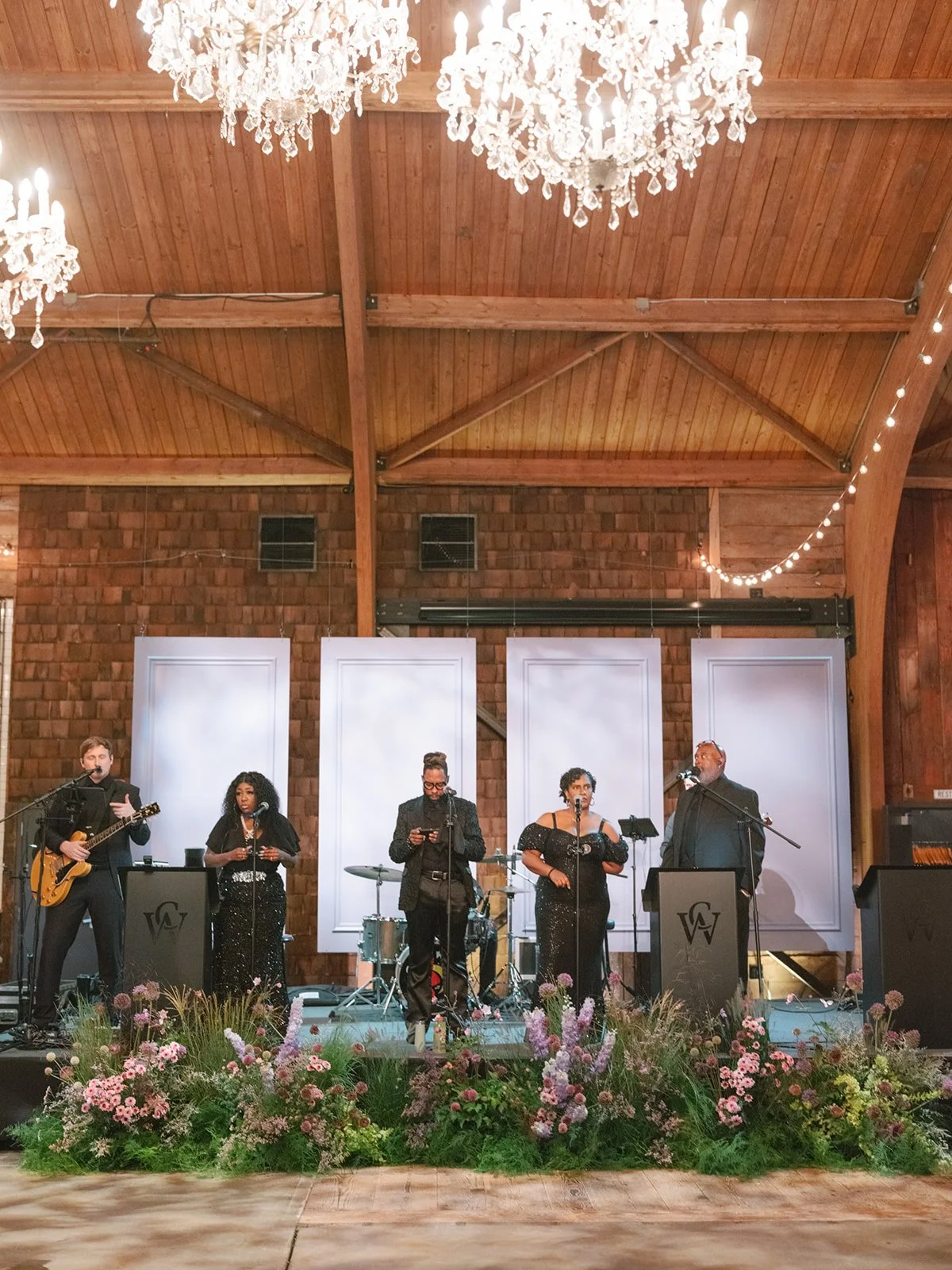 A musical band performing on a stage decorated with flowers, inside a venue with wooden ceiling beams and chandeliers.