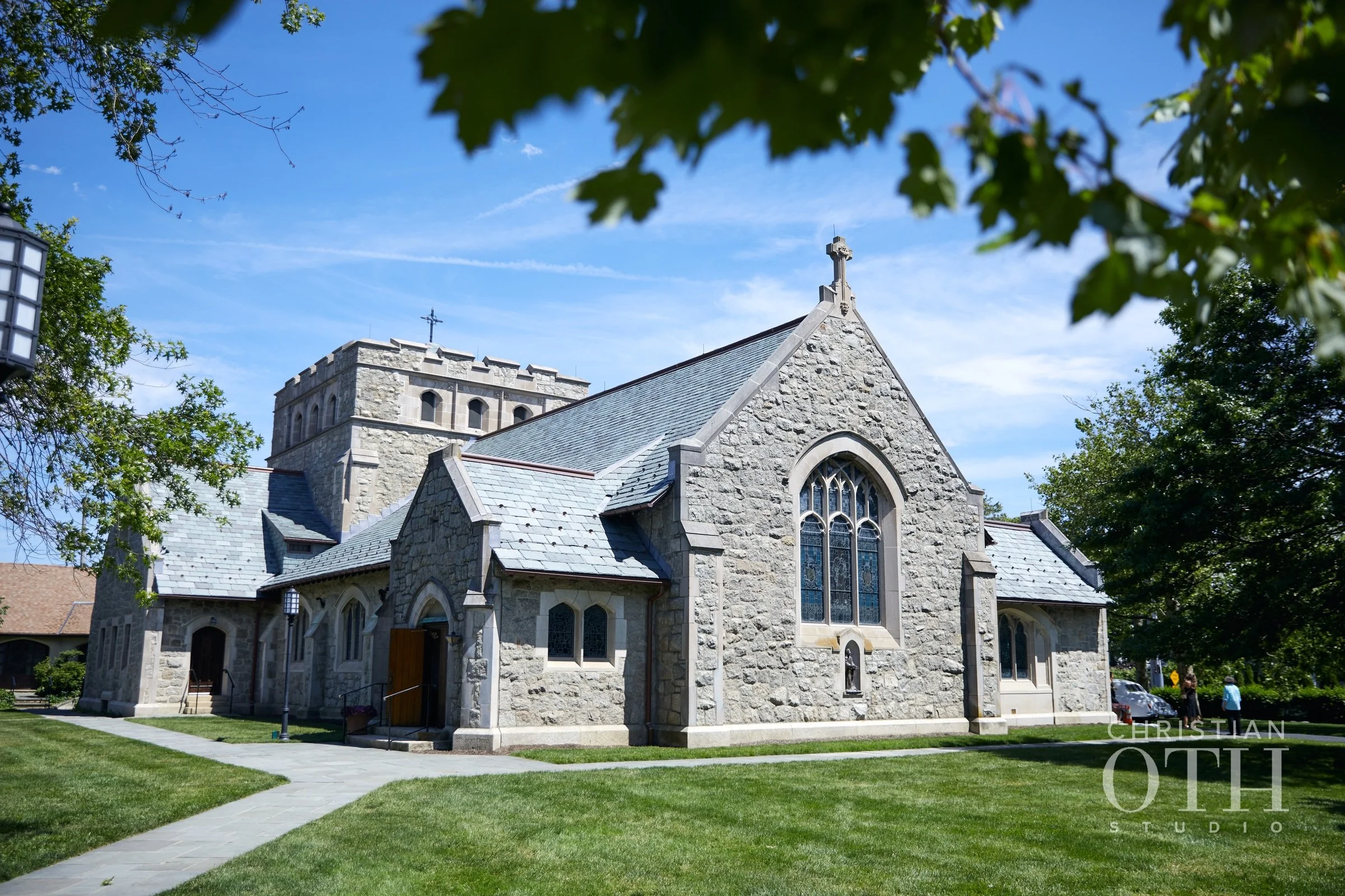Stone church with arched stained glass windows, a sloped roof, and a small cross on top. Surrounded by green grass and trees, with people walking nearby under a partly cloudy blue sky.