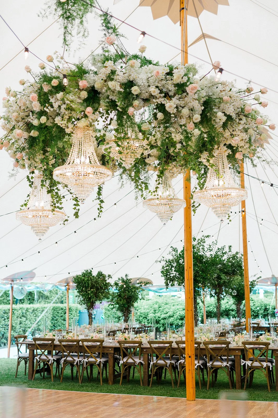 Elegant outdoor event tent decorated with a floral centerpiece featuring white and pink flowers with chandeliers hanging from a lush floral arrangement, and a long table set with place settings and floral accents.