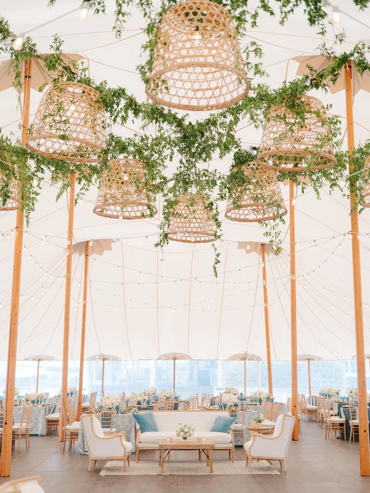 Elegant event tent decorated with hanging wicker lanterns and greenery, featuring a cozy seating area with white sofas, armchairs, and tables, alongside guest tables with floral centerpieces and umbrellas.