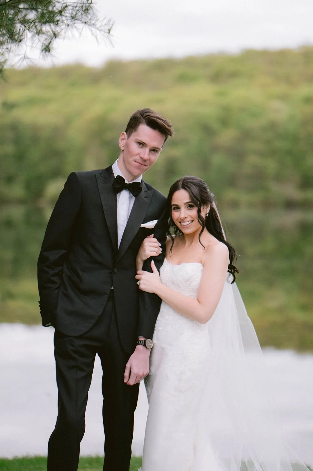 A couple dressed in wedding attire standing outdoors near a body of water, with greenery in the background. The groom is in a black tuxedo, and the bride is in a white wedding gown.
