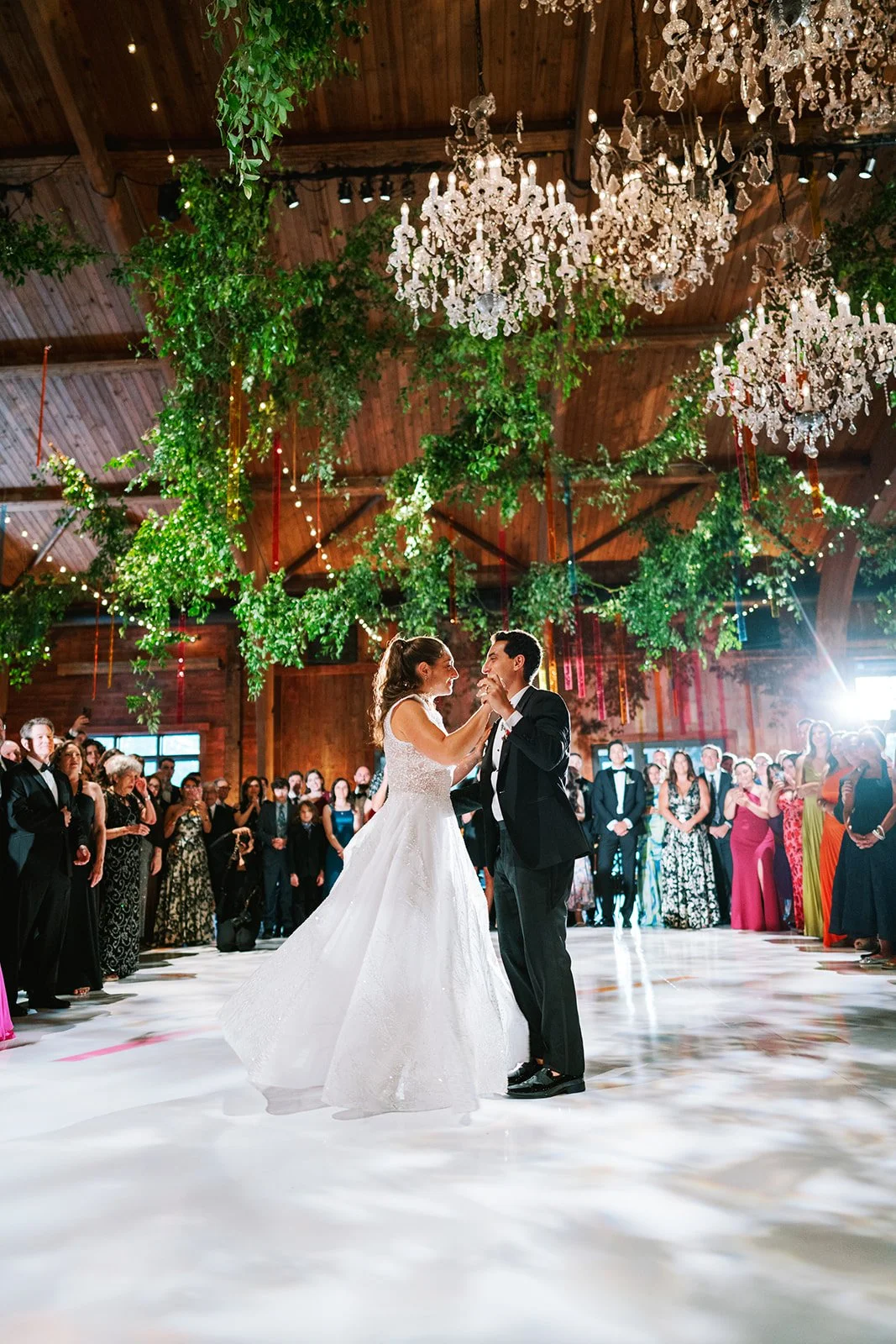 Bride and groom dancing at their wedding reception with guests watching in the background, decorated with chandeliers and greenery.