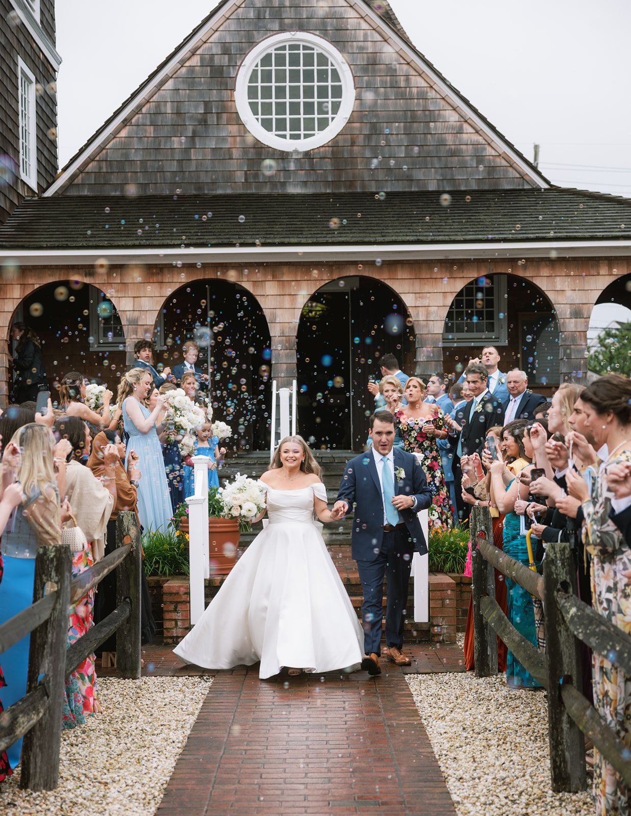 A newlywed couple walks down the aisle outside a church, surrounded by guests throwing confetti and taking photos. The bride wears a white wedding dress and holds a bouquet, while the groom wears a navy suit with a light blue tie. The church has a ru