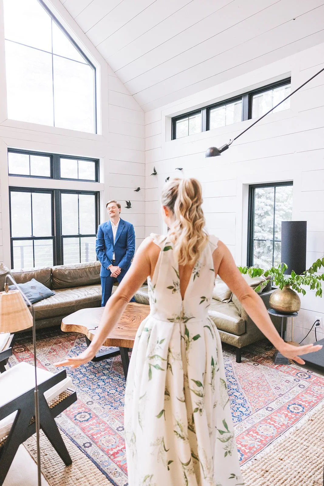 A woman in a white floral dress with a deep V-back is facing a man in a blue suit in a bright, modern living room with large windows, white paneled walls, a brown leather sofa, a wooden coffee table, and decorative plants.